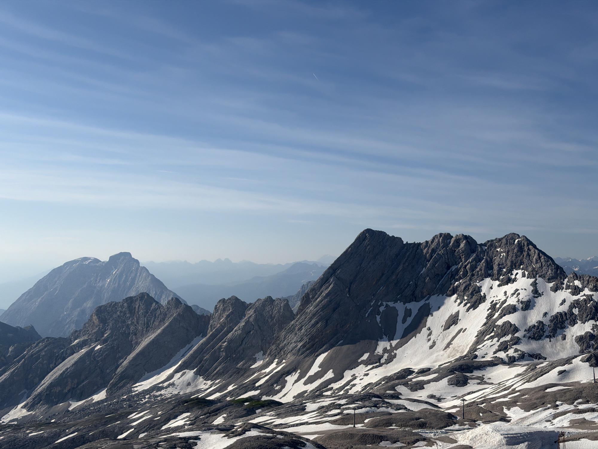 This is a wide-angle, high-altitude photograph taken from the Zugspitze, Germany's highest peak, near Garmisch-Partenkirchen. The image showcases a dramatic mountain landscape under a clear blue sky with thin, horizontal clouds. In the foreground, rugged, grey, rocky slopes are sparsely covered with patches of white snow. Several utility poles or markers are visible in the lower portion of the frame. The middle ground is dominated by a jagged mountain range with steep, shadowed faces and snow-dusted ridges. In the background, several layers of distant mountains fade into a hazy, blue atmosphere, suggesting a vast and expansive view. The overall impression is one of a serene, majestic, and remote alpine environment. There are no people or animals visible in the image.