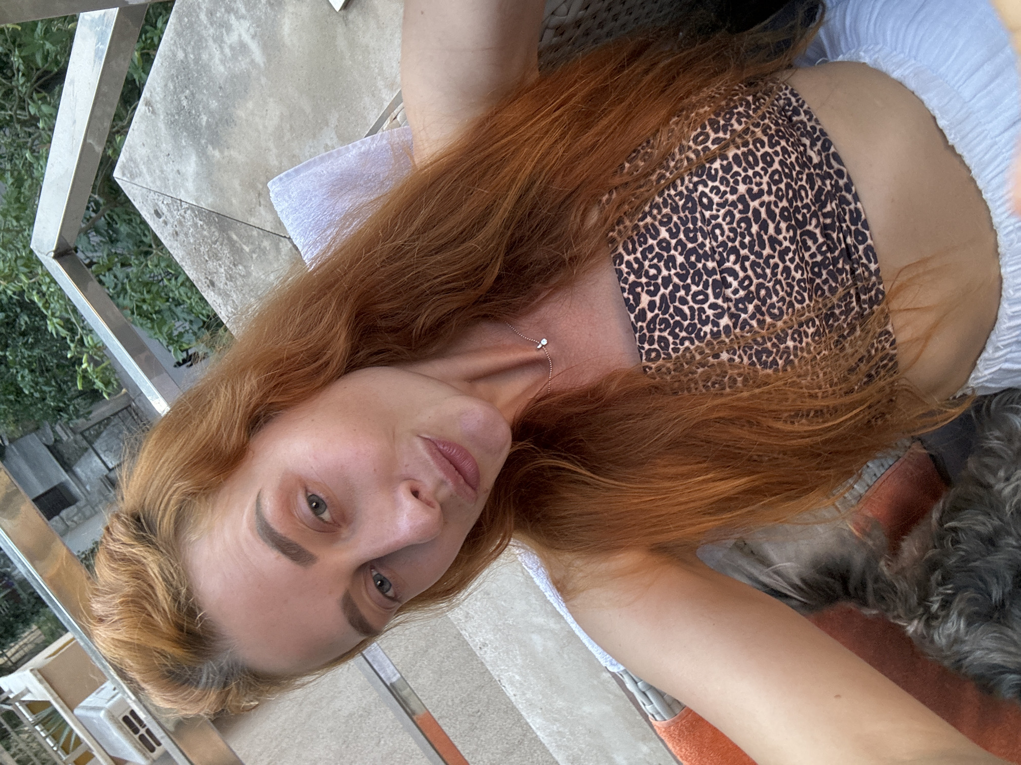 In Budva, Montenegro, a woman with long, auburn hair poses for a selfie. She is wearing a leopard-print bandeau top and white pants, seated outdoors on a woven chair. Her hair is styled loosely, with some strands falling forward. She has a subtle smile and her eyes are looking towards the camera. A small, delicate necklace with a diamond pendant adorns her neck. In the background, a railing and lush green foliage are visible, suggesting an outdoor setting, possibly a balcony or patio. To her left, an orange towel is draped over the chair. Part of a small, dark-furred dog is visible in the lower left corner of the image, resting against her. The scene appears to be bathed in natural light.