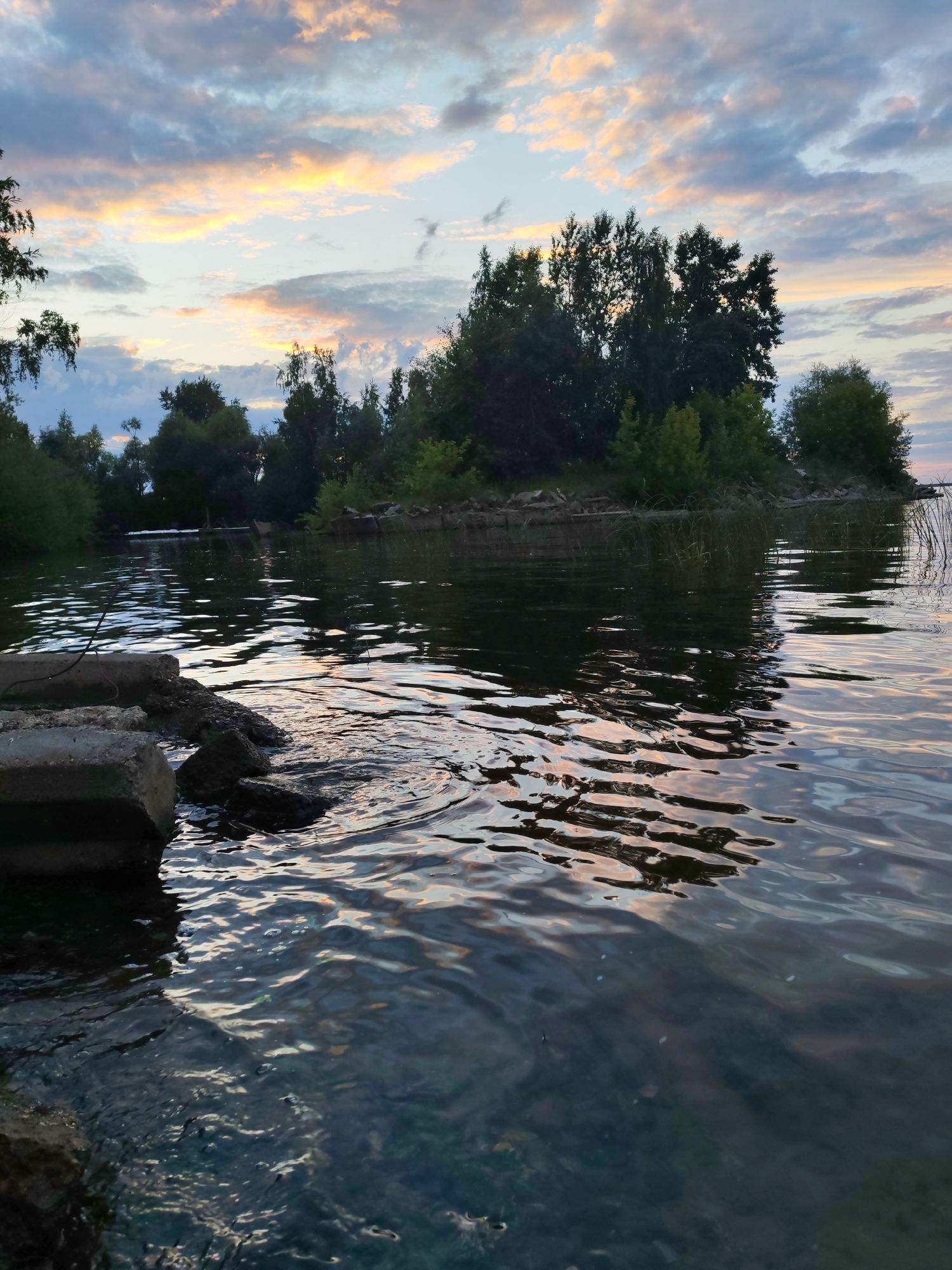 The image depicts a tranquil scene at the Kama River in Chaikovsky, Russia, during sunset. The sky is adorned with a dramatic display of clouds, painted in hues of orange, yellow, and blue, creating a soft, diffused light. A dense line of trees and foliage forms the backdrop, bordering the river and creating a natural, untouched landscape. In the foreground, concrete debris rests on the riverbank, partially submerged in the calm water, suggesting a former man-made structure or remnants of construction. The water's surface is gently rippled, reflecting the colors of the sunset and the surrounding greenery. The overall atmosphere is serene and picturesque, capturing the beauty of nature at dusk.
