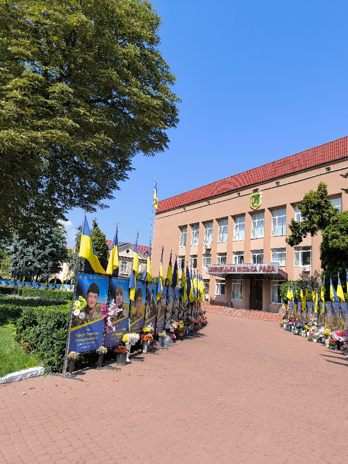 An outdoor, eye-level shot captures the Illintsi City Council (ІЛЛІНЕЦЬКА МІСЬКА РАДА) building in Illintsi, Ukraine, under a clear blue sky. The building features a light peach-colored facade, multiple windows, a red tiled roof, and the city's coat of arms above the entrance. A wide, reddish-orange paved pathway leads directly to the building's entrance.
Flanking both sides of this pathway is a double-row arrangement of vertical memorial banners. Each banner displays a portrait photograph, primarily of men in military attire, with Ukrainian text below the image. A Ukrainian national flag (blue and yellow) is attached to the top of each banner. At the base of each banner, various floral arrangements, consisting of both fresh and artificial flowers in buckets and vases, are positioned on the ground.
To the left of the pathway, a large, mature tree with dense green foliage is prominent, along with manicured green bushes and several evergreen trees. The scene is brightly illuminated by direct sunlight.