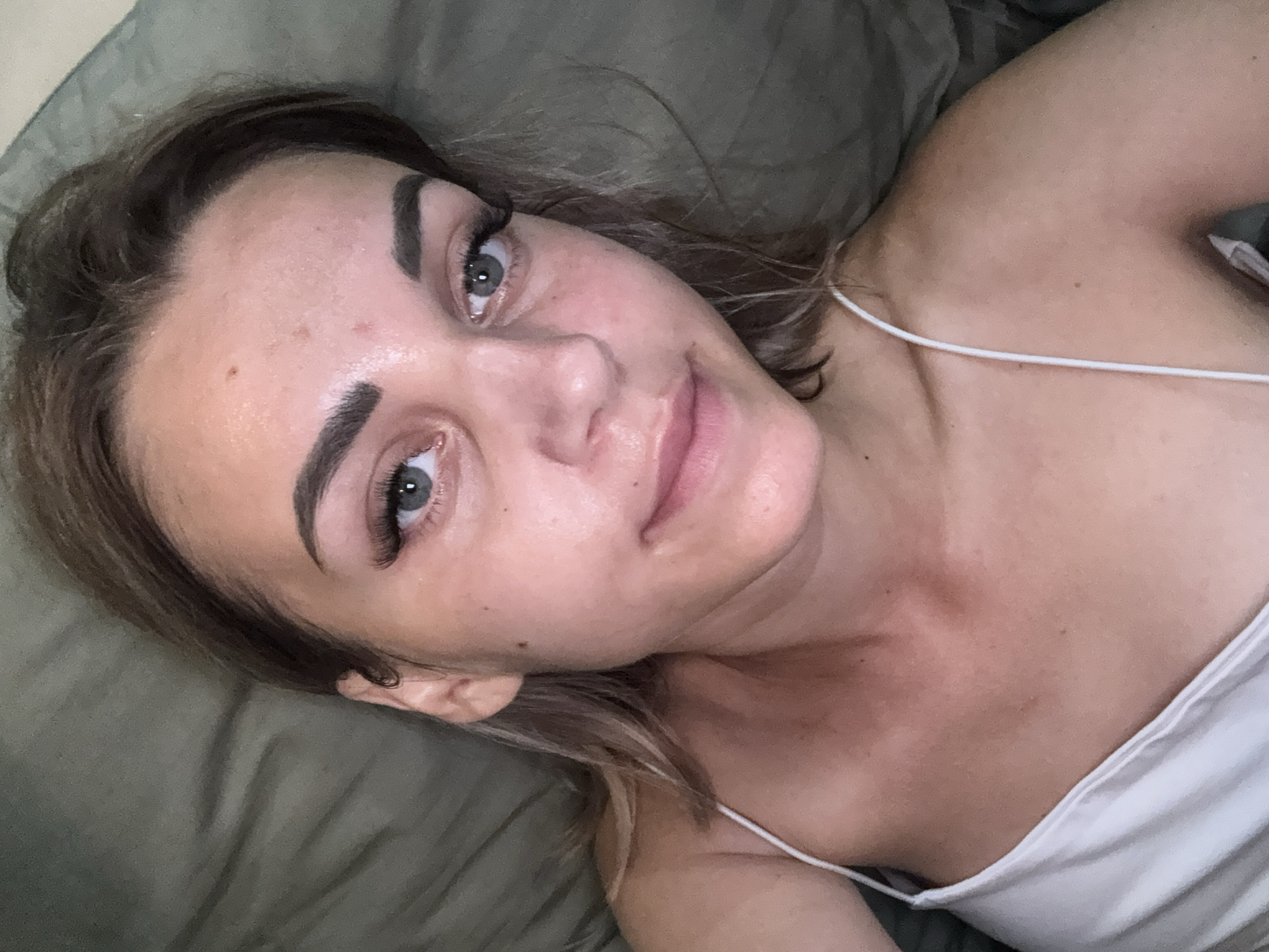 A close-up, high-angle selfie shows a young woman lying on a dark green textured surface, likely a bed or couch. Her head is tilted slightly to her left, and she is looking directly at the camera with a faint smile. She has fair skin with some redness on her cheeks and forehead, and a few small blemishes. Her eyebrows are well-defined and dark, and she is wearing false eyelashes. Her eyes are light blue-gray. Her hair is light brown and is slightly messy, framing her face. She is wearing a white tank top with thin spaghetti straps. The background is out of focus, showing only the texture of the surface she is lying on. There is no indication of the city of Yuzhno-Sakhalinsk, Russia, or any other specific location. The scene is intimate and relaxed.