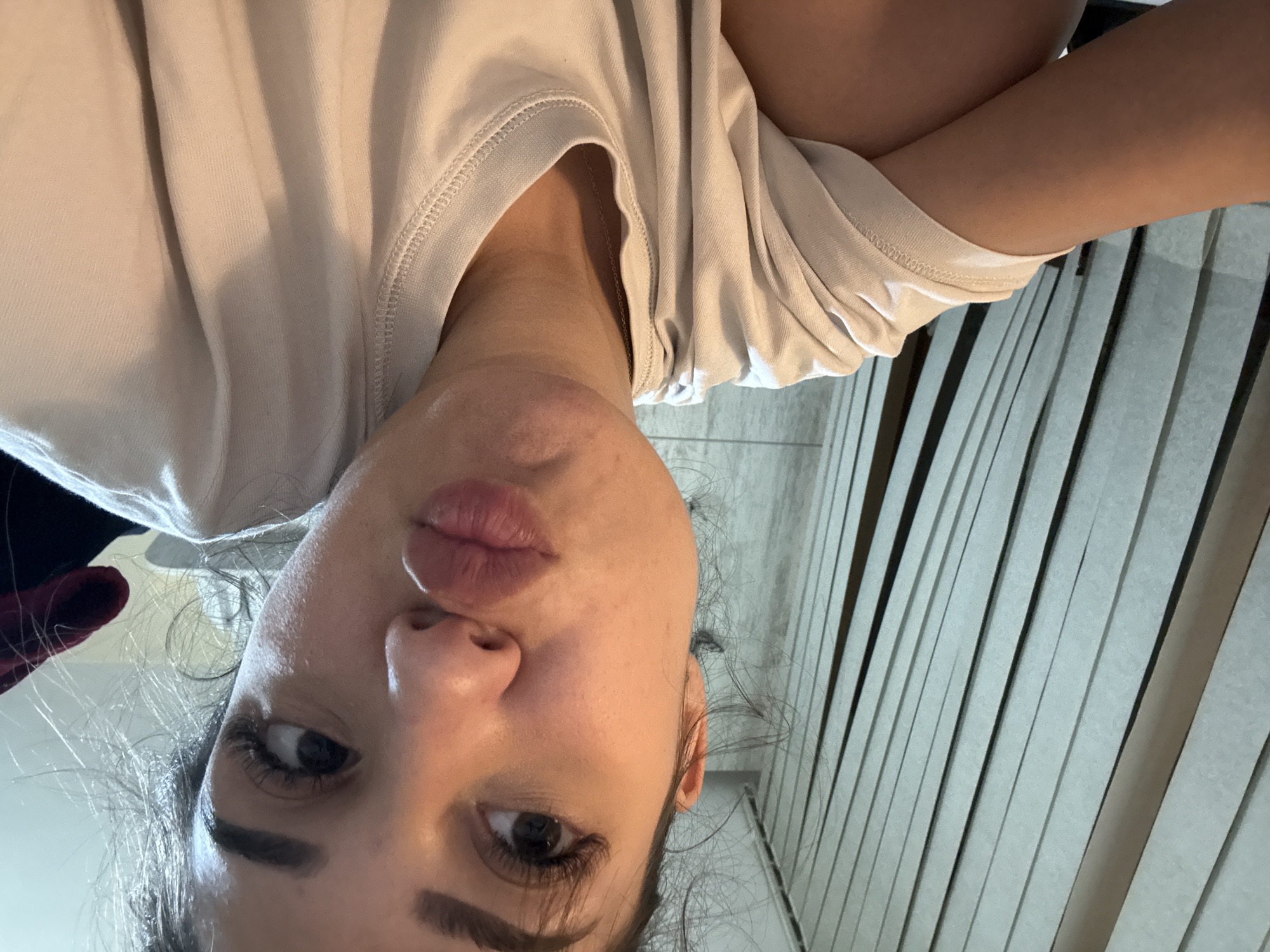 The image shows a close-up, selfie-style shot of a young woman with her mouth puckered. She is wearing a light-colored t-shirt. The background consists of vertical blinds, suggesting an indoor setting, possibly a home or hotel room. There is no indication of the location being Plovdiv, Bulgaria.