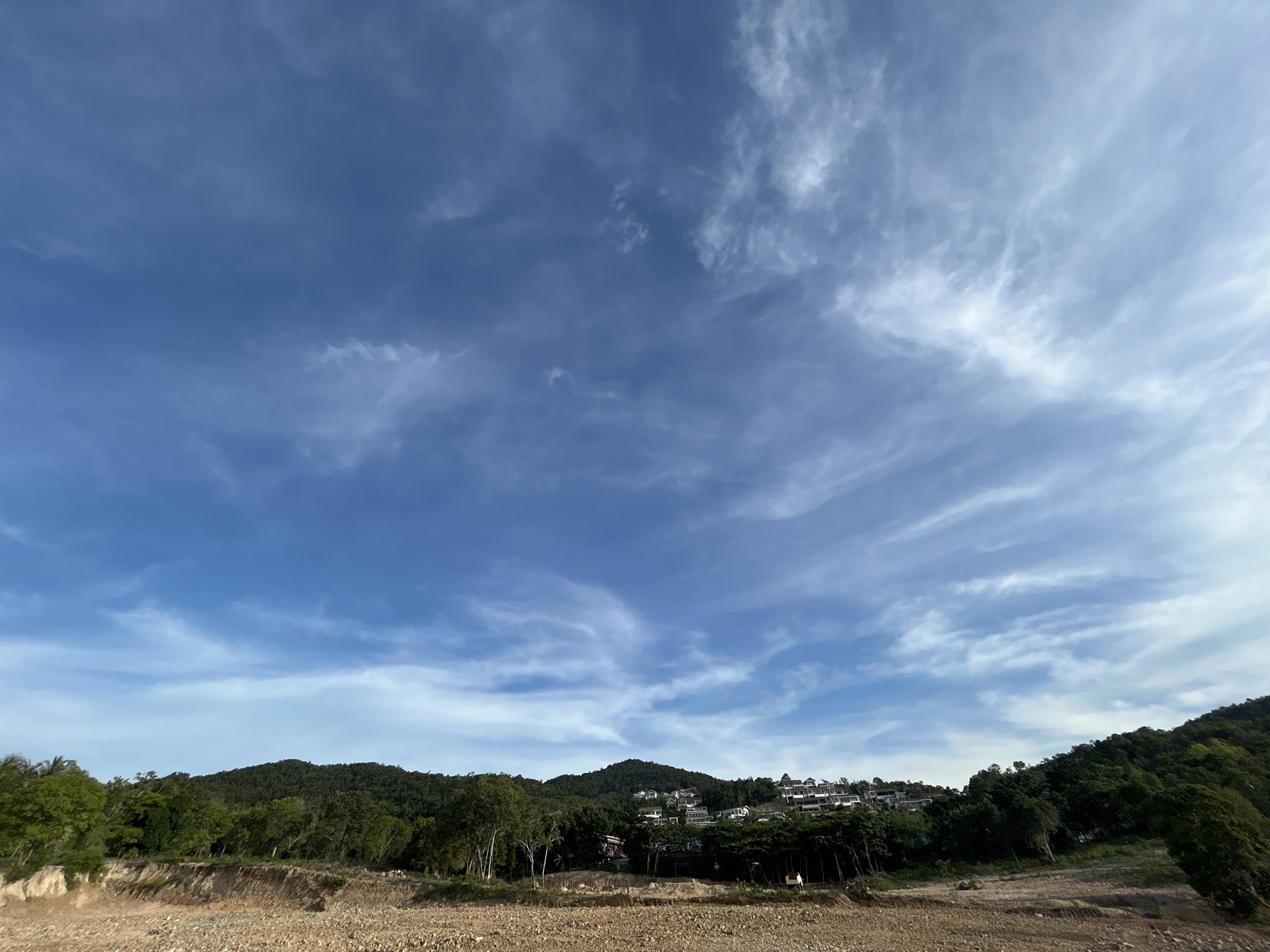 The image depicts a landscape in Baan Wang Ta Kien, Thailand, under a partly cloudy blue sky. The ground is a barren, earthy brown, with evidence of excavation. A line of trees separates the foreground from a hillside densely covered with lush green vegetation. Residences are visible on the hillside, nestled among the trees. The sky is mostly blue with wispy, cirrus-like clouds spread throughout.