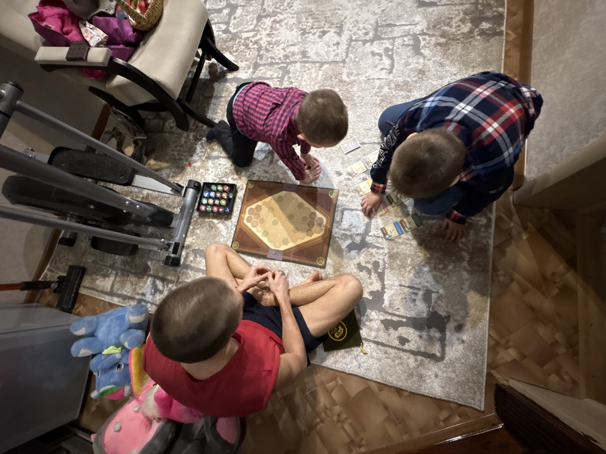 **Location:** Indoors, possibly a residence in Ulyanovsk, Russia, based on the presence of typical Russian household items and the general style of the interior.
**People:**
* Three individuals, likely children or young adolescents.
* One individual is wearing a red shirt and dark shorts, sitting cross-legged near the bottom of the image.
* Another individual is wearing a red and black plaid shirt and dark pants, kneeling on the left side of the board game.
* The third individual is wearing a blue and red plaid shirt and dark pants, kneeling on the right side of the board game.
**Subjects/Objects:**
* Board game with a hexagonal tile pattern on the playing surface, surrounded by cards.
* A set of colored game pieces next to the board game.
* A rug with a neutral color palette.
* A chair and other household furniture visible in the background.
* Exercise equipment, specifically an elliptical machine, is visible on the left side of the image.
* Various stuffed animals are visible on the left side of the image.
**Actions/Interactions:**
* The three individuals appear to be engaged in playing the board game.
* They are positioned around the board game, presumably taking turns or strategizing.
**Environment:**
* The scene is indoors, possibly a living room or playroom.
* The lighting appears to be artificial, likely from overhead fixtures.
* The overall impression is of a domestic scene with children playing a game.