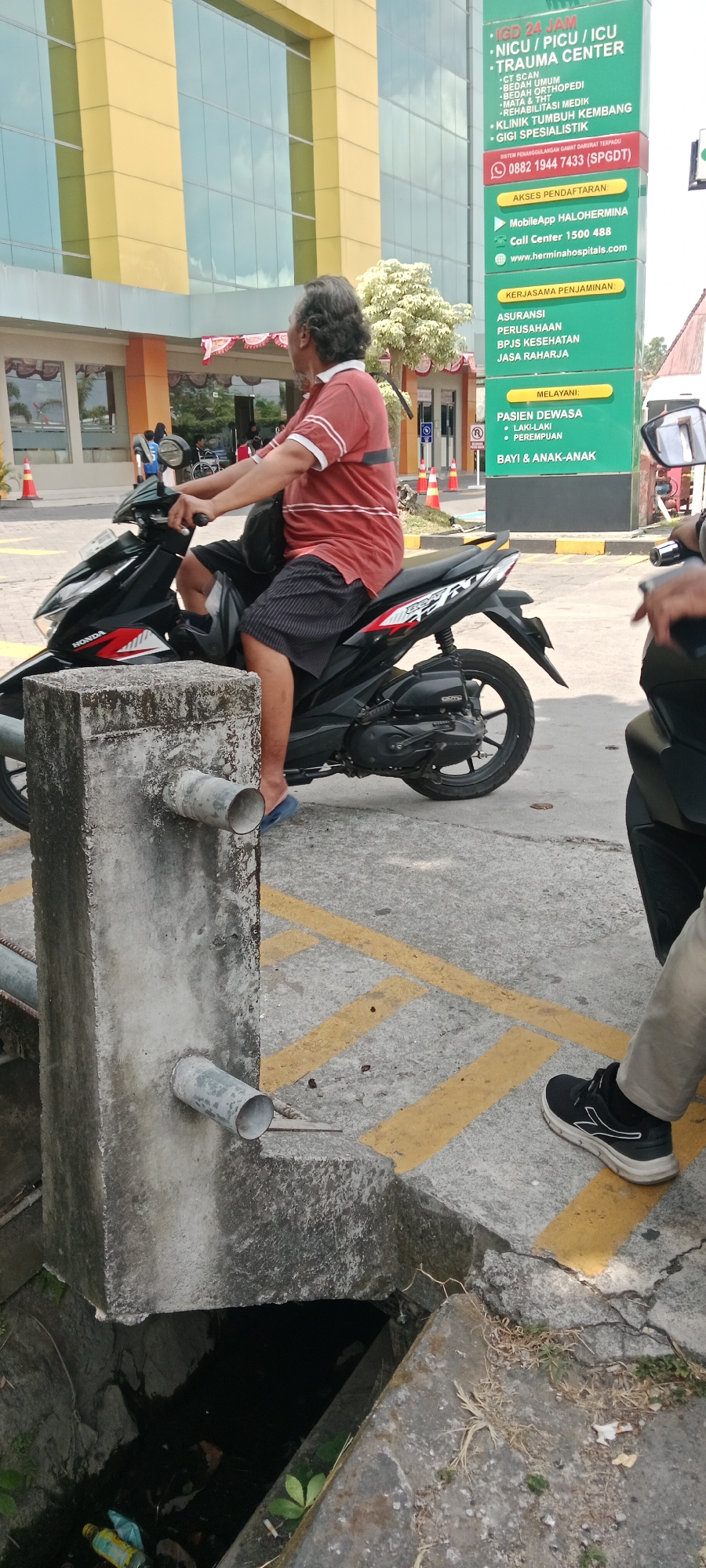 In Maguwoharjo, Indonesia, a man with long, gray hair and a beard is seated on a black Honda motorcycle. He is wearing a reddish-orange polo shirt with white stripes on the collar and sleeves, and dark-striped shorts. His left foot is clad in a blue sandal, while his right foot is not visible. The motorcycle is positioned next to a weathered concrete pillar that has two horizontal metal pipes protruding from it. The pillar is situated on a concrete surface marked with yellow lines. In the background, a modern building with a yellow facade and large glass windows stands prominently. To the right of the building, a tall green sign displays various services offered by a medical facility, including "IGD 24 JAM, NICU / PICU / ICU, TRAUMA CENTER," and contact information. Another person's legs are visible on the far right of the frame, wearing light-colored pants and a black athletic shoe with white accents. The environment appears to be an outdoor area, possibly a hospital or clinic entrance, with some vegetation and scattered debris on the ground.