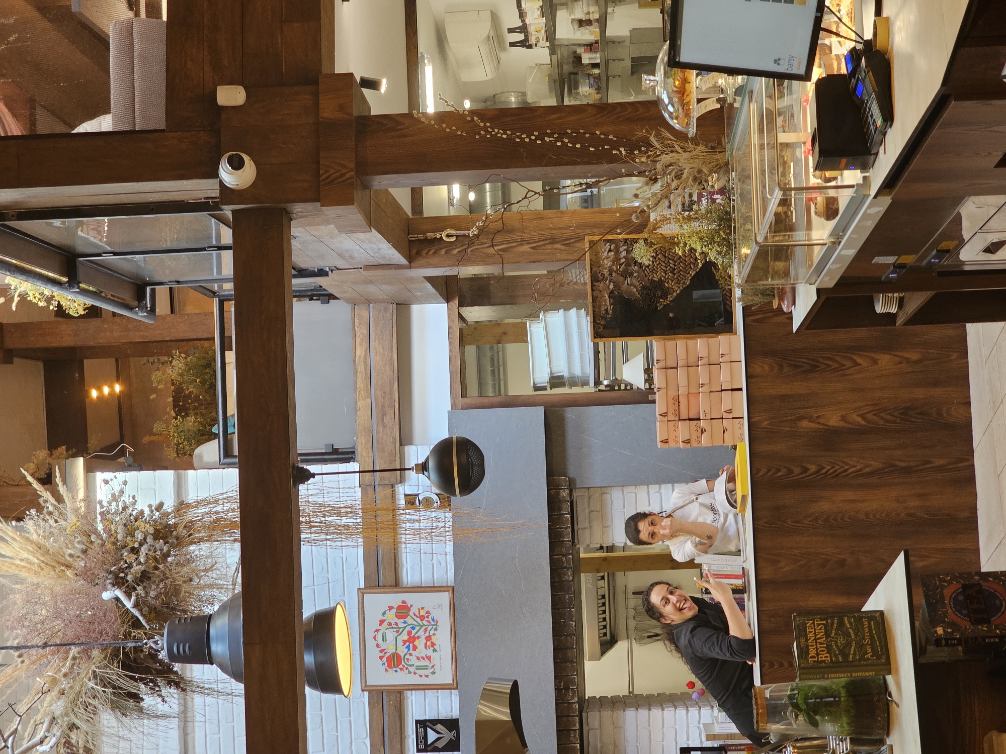 The image depicts the interior of a cafe or restaurant with a rustic, natural aesthetic. Two women are visible behind a wooden counter. The woman on the left, wearing a black top, is leaning forward, sticking out her tongue and making a peace sign with her fingers. The woman on the right, wearing a white apron over a white top, is looking towards the viewer and has her fingers near her nose.
The counter is made of wood and is lined with various items, including books, a terrarium, and a display case with desserts. Behind the counter, there are shelves with more items, and a kitchen area with white brick walls. The cafe's interior features exposed wooden beams, hanging plants, and decorative dried floral arrangements. There is a security camera mounted on a wooden beam. In the background, a monitor displays the word "barsy" which is likely the name of the establishment. The overall ambiance is warm and inviting, with natural materials and soft lighting.
The location is identified as Plovdiv, Bulgaria, implying this scene is within a specific establishment in that city.