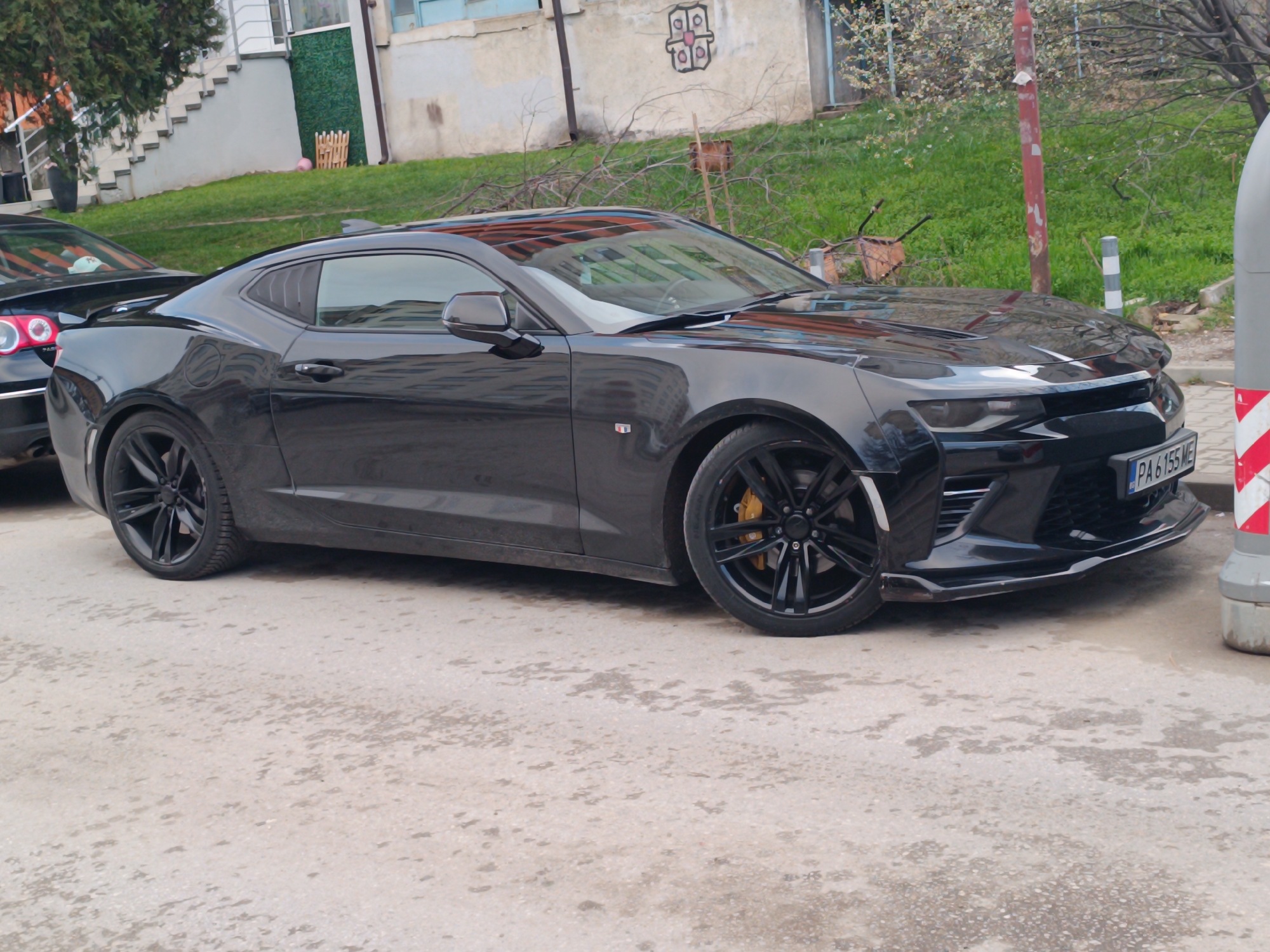 The image depicts a black Chevrolet Camaro parked on a street in what appears to be a residential area. The car is oriented towards the right of the frame, with its passenger side visible. A black car, likely a Volkswagen Passat based on the tail lights, is partially visible in the background behind the Camaro.
The Camaro has black rims and yellow brake calipers, and a Bulgarian license plate "PA 6155 ME" is visible on the front. The car appears to be a performance model based on the front splitter.
The surroundings include a mix of buildings, trees, and greenery. A house with a staircase leading to the entrance is seen in the background. A pole with red and white striped markings stands near the car's front bumper. The ground is paved and shows signs of wear.
Based on the license plate and general appearance of the environment, the location is likely in Sofia, Bulgaria.