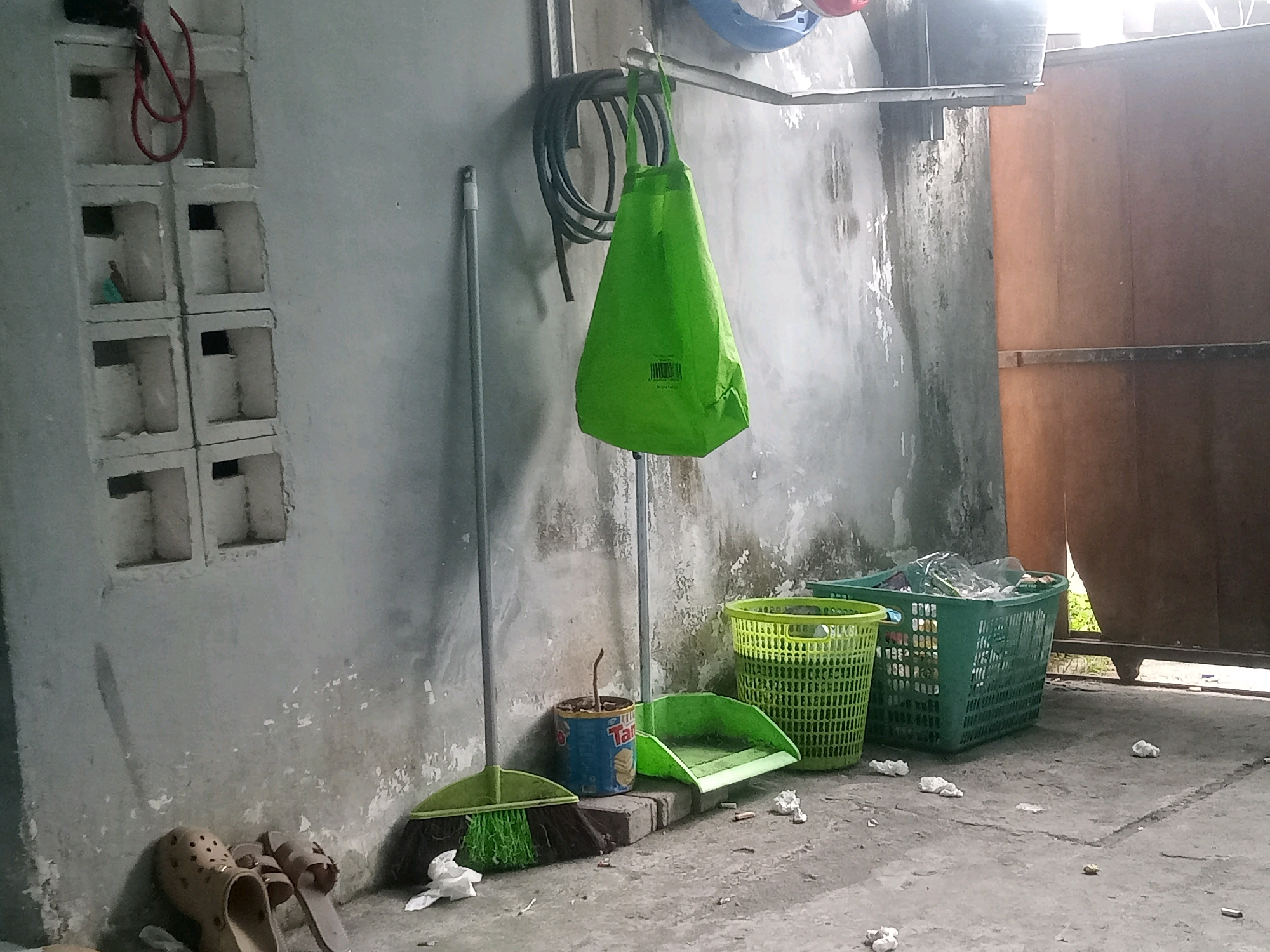 The image depicts a cluttered storage area or backyard in Ranjok, Indonesia. In the foreground, a broom with a green head and a metal handle stands upright next to a small, rusty can with a plant growing in it. A green dustpan rests on the ground in front of the can. To the right, two green plastic baskets are visible; one is empty, and the other contains plastic bags and other debris. A pair of brown sandals are placed against a rough, grey concrete wall on the left side of the frame. Above the sandals, the wall has a series of rectangular openings. A large, bright green fabric bag hangs from a metal bar on the wall, with a coiled black hose behind it. The ground is concrete, with scattered cigarette butts and crumpled white paper. In the background, a tall, wooden door or partition is visible. The overall impression is one of an unkempt, utilitarian space.
