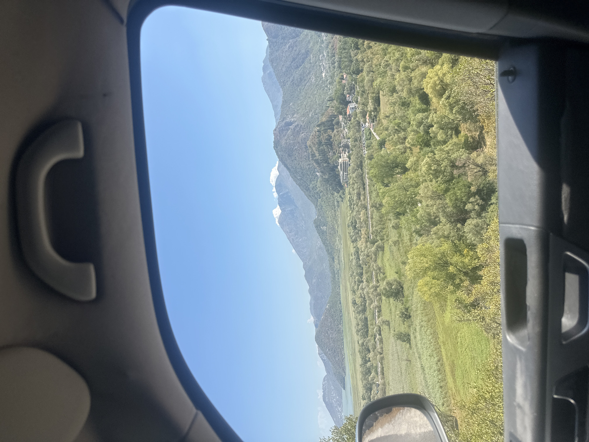 The image, taken from inside a vehicle, offers a scenic view of a mountainous landscape near Braćeni, Montenegro. The foreground features the interior of the car, including the gray door panel with visible handles and a black interior trim. The side mirror is visible on the left, reflecting a narrow, dirt road.
Beyond the car's window, a vast expanse of green fields and trees dominates the midground. A body of water, likely a lake, is visible to the left, reflecting the clear blue sky. In the distance, imposing mountains rise, their slopes covered in lush vegetation. Some of the higher peaks are dusted with snow, contrasting with the green below. A small cluster of buildings, possibly a village or resort, is nestled amidst the trees on a lower mountain slope. The sky is a bright, cloudless blue, indicating a sunny day. The overall impression is one of natural beauty and tranquility.