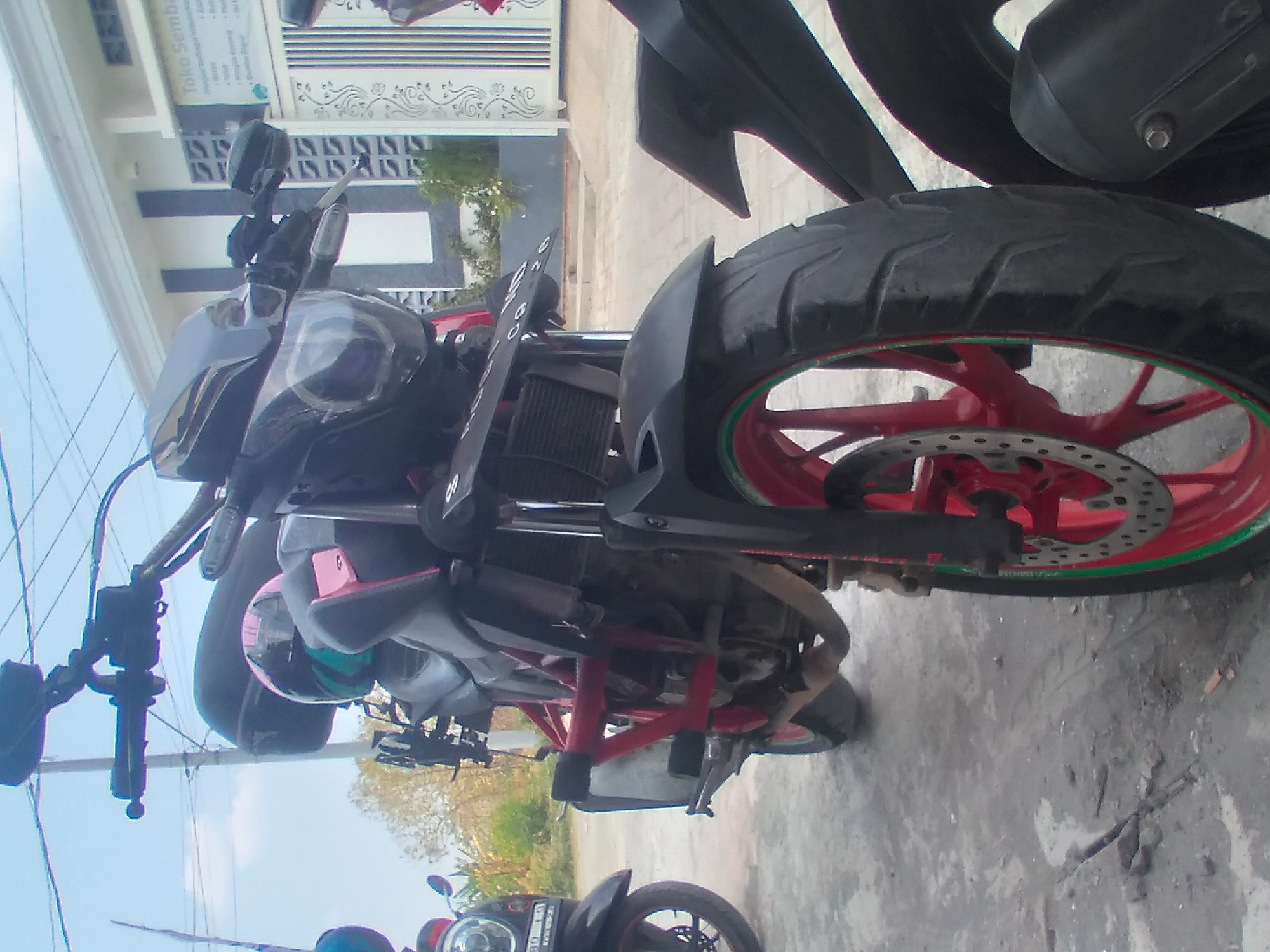 The image shows two motorcycles parked on a paved surface in what appears to be a residential or commercial area. The motorcycle in the foreground is black and red, with a visible engine, exhaust pipe, and red rims on the wheels. A pink and teal helmet is placed on the seat of this motorcycle. The second motorcycle, partially visible on the left, is black with a circular headlight. In the background, a building with white and dark grey accents and decorative gates is visible. A sign on the building reads "Toko Semb". The environment suggests a sunny day, with a blue sky and some trees visible in the distance. The location is identified as Sumberjo, Indonesia. There are no people visible in the image. The scene is static, depicting parked vehicles in an outdoor setting.