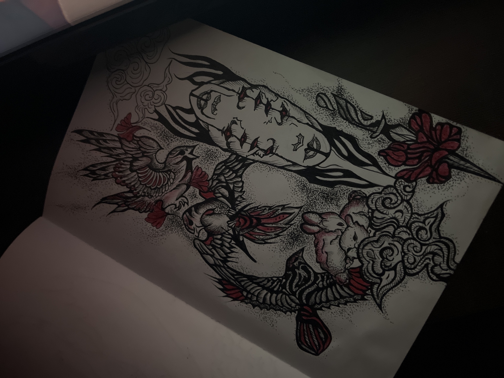 This media file presents a detailed pen and ink drawing on a white sketchbook page, photographed in low light. The intricate artwork, reminiscent of neo-traditional or Japanese tattoo art, features a complex composition of symbolic elements rendered in black ink with selective red accents and extensive stippling for shading.
Subjects within the drawing include a stylized, mask-like face or figure with multiple eyes and expressive features, positioned horizontally across the upper middle section. Swirling lines and dotted shading emanate from around this central face. Below and to the left, two ornate bird-like creatures, possibly phoenixes or mythical birds, are depicted in dynamic poses with detailed feathers, some highlighted in red. To the right and bottom, an intricate creature resembling a rabbit or a similar animal is intertwined with elaborate cloud-like patterns, also featuring red details. A dagger with a red floral hilt and wavy blade elements is visible in the upper right.
The scene is an open sketchbook displaying this finished drawing. The setting appears to be indoors, indicated by the dim lighting and shadows cast across the page, particularly on the left and bottom margins, suggesting it might be evening or night. The primary activity is the artwork itself, a static display of the completed drawing.
Notable details include the striking contrast of black ink and vibrant red against the white paper, and the consistent use of stippling for texture and depth throughout the composition. The elements are interconnected, creating a flowing and visually engaging design. There is no visible text in the image.