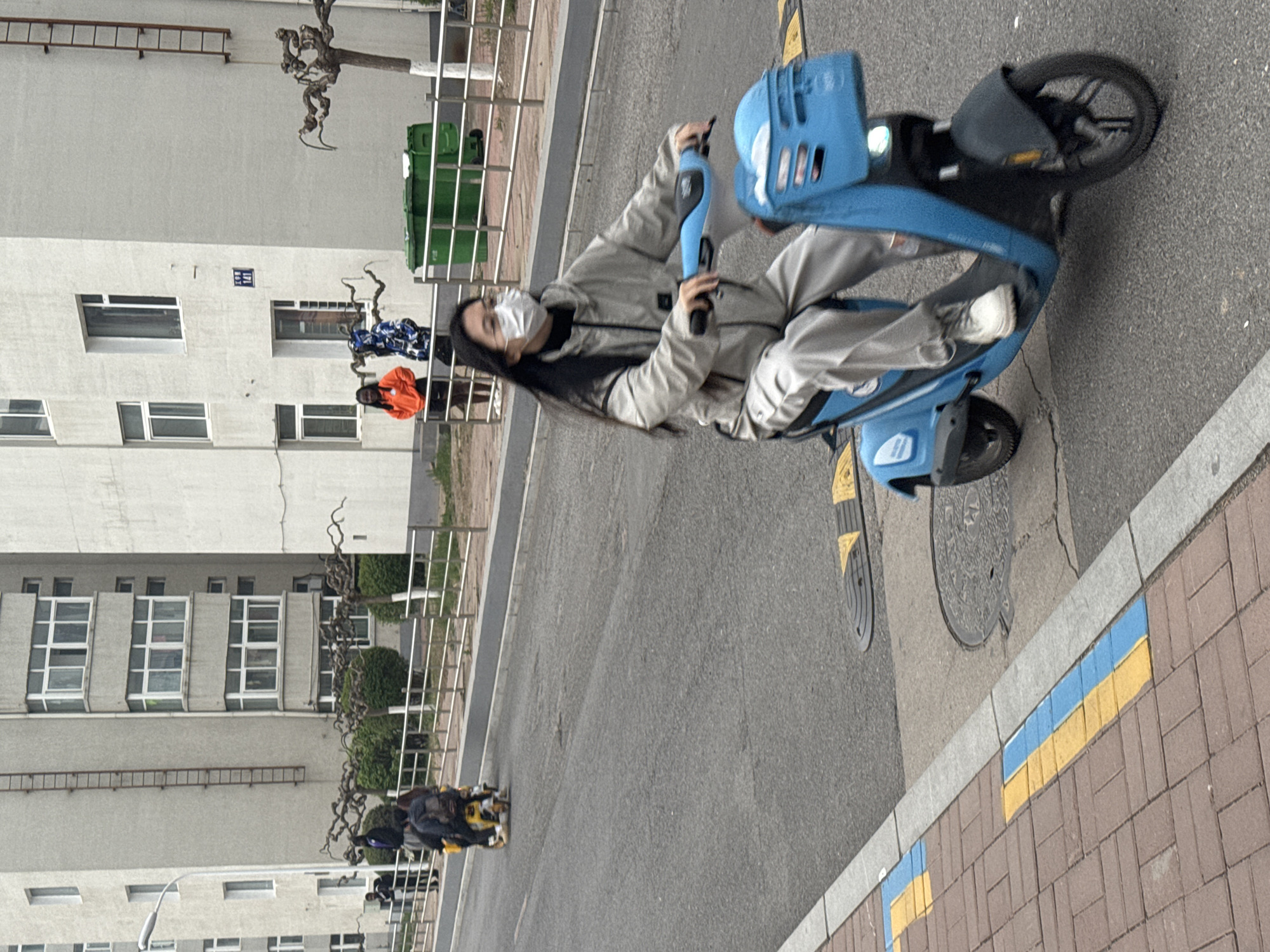 The image depicts a daytime street scene in an urban or suburban area, identified as Zhongtun, China. The primary subject is a young woman, positioned centrally, riding a blue electric scooter. She is dressed in a light gray jacket, light gray trousers, and white sneakers, with long dark hair, and is wearing a white face mask. She appears to be stationary or moving slowly, looking towards the camera. The scooter is bright blue, featuring a front basket and integrated lights.
In the background, several light-colored, multi-story apartment buildings with numerous windows line the street. A metal ladder leans against one of these buildings, and a small blue sign with white numbers, possibly "117", is visible on another. Behind a metal fence, two green trash bins are visible, along with a gnarled, leafless tree. Further down the street, other parked motorcycles or scooters can be seen.
The street surface is asphalt, bordered by a brick sidewalk. The curb separating the street from the sidewalk features distinct painted stripes in segments of blue and yellow. A manhole cover is visible on the asphalt near the sidewalk. The lighting suggests a clear day. The scene captures a moment of daily life, with the woman on the scooter being the focal point of an otherwise calm and routine setting.