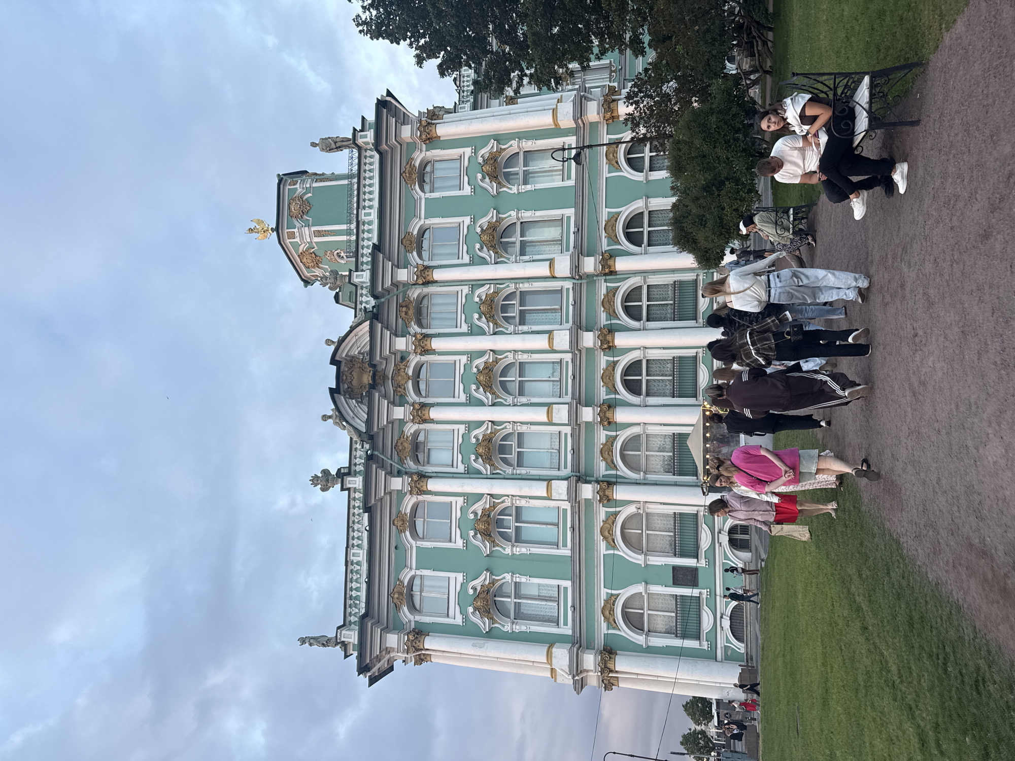 The image shows the exterior of the Winter Palace, a former imperial palace and now part of the State Hermitage Museum, located on Palace Square in Saint Petersburg, Russia. The palace is a grand, historic building with a distinctive green facade and white architectural details. Several people are seen walking on a gravel path in front of the palace and sitting on benches in a grassy area. The sky is overcast, suggesting it is either early morning or late evening. The overall scene depicts a popular tourist destination with visitors enjoying the architecture and grounds.