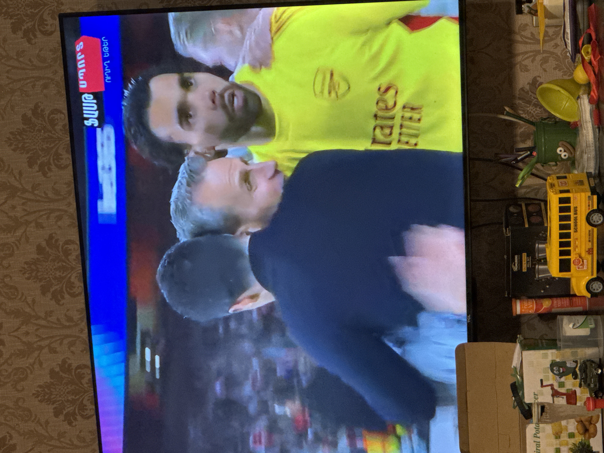 The image shows a television screen displaying a scene of two men in what appears to be a sports celebration, superimposed on a messy table with various items.
On the screen, one man is wearing a yellow sports jersey with the word "Emirates" on it, likely indicating the team he represents. He is being embraced by a second man wearing a navy blue jacket. The background of the screen indicates a crowd of people, suggesting a sports event. The screen also shows Armenian text indicating the sport event is live, possibly filmed in the city of Yerevan, Armenia.
On the table next to the TV, there is a yellow school bus toy, a green toy with googly eyes, a box of Spiral Potato Slicer, and various other items cluttering the space. There appear to be pens, tools, containers, and other small objects scattered haphazardly.