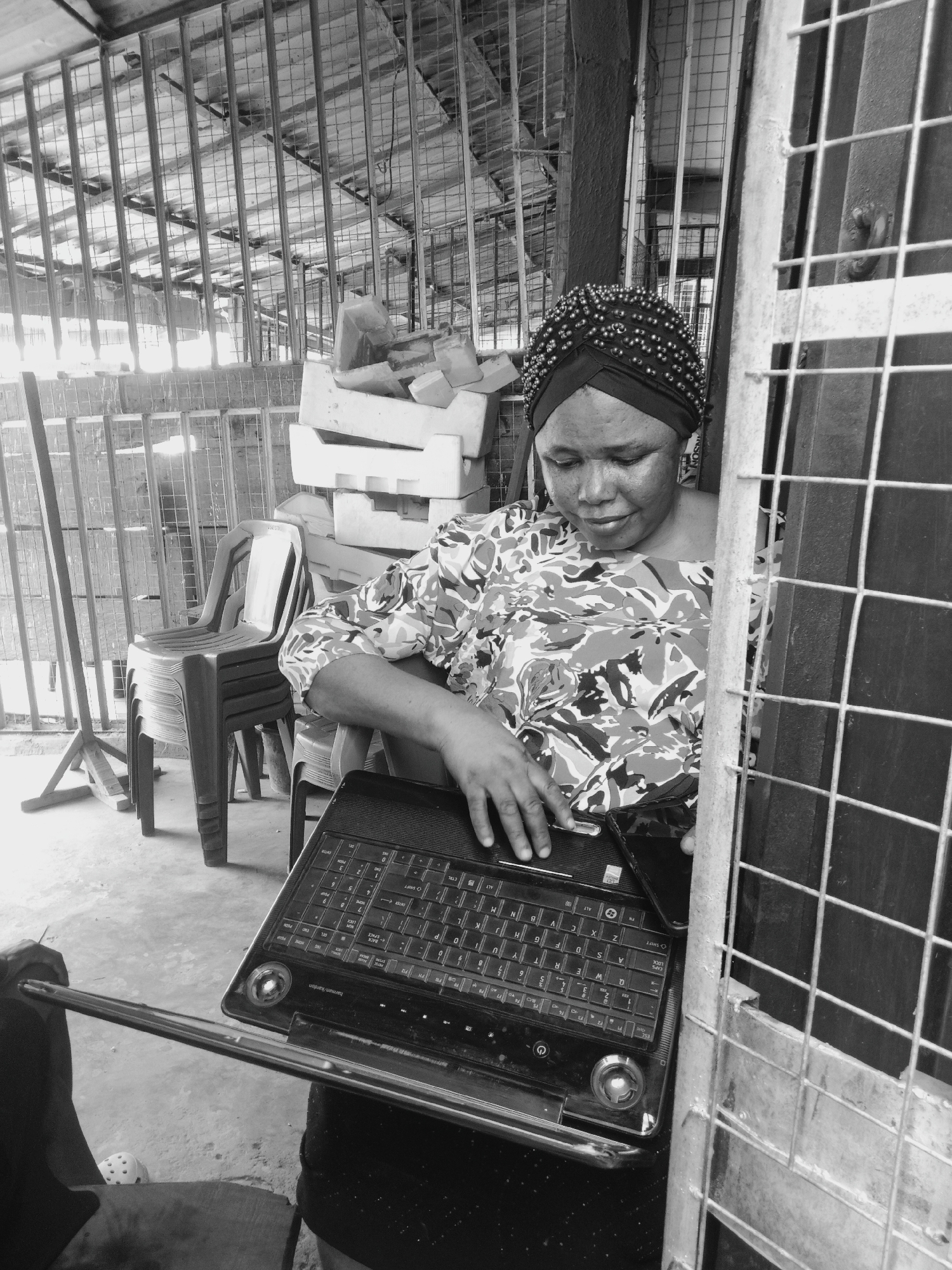 This black and white image captures a woman in Onitsha, Nigeria, intently focused on a laptop in a functional, semi-enclosed setting. The woman, centered in the frame, wears a dark, beaded headwrap or turban and a patterned, likely floral or abstract, long-sleeved top. Her gaze is directed downwards at an open black laptop resting on her lap, with her right hand positioned over the keyboard as if typing or navigating. A smartphone is also visible, placed next to the laptop.
The background of the scene is characterized by a lattice of metal grates or cages, suggesting an industrial or commercial environment such as a market stall, warehouse, or workshop. To the left, a stack of dark-colored plastic chairs is visible, along with several irregularly shaped white objects that appear to be styrofoam blocks or containers. Another section of metal grating, possibly part of a gate or barrier, occupies the right side of the foreground, partially framing the woman.
The monochromatic treatment emphasizes the textures of the metalwork, the woman's headwrap, and the details of the laptop. Bright, possibly natural, light illuminates the scene, indicating it is daytime. The laptop clearly displays "harman/kardon" branding, along with various keyboard keys such as "SHIFT," "LOCK," "Q," "W," "E," "Z," "S," "X," "V," "5," and "9." The woman's expression is one of calm concentration, suggesting she is engaged in a task or activity requiring her attention on the digital device.