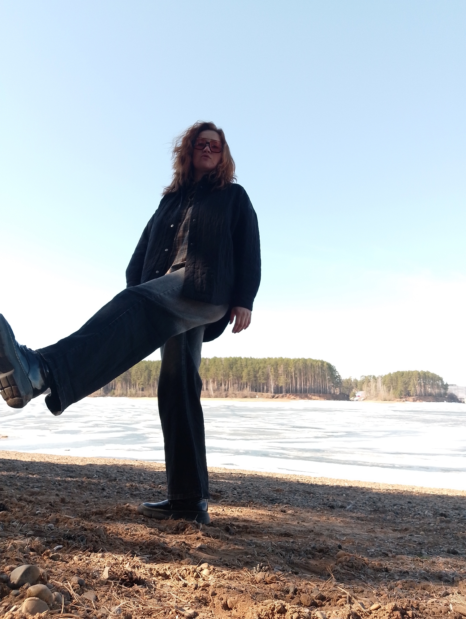 A person stands on a pebbly shore, with one leg extended forward. They are wearing dark-colored jeans, shoes, a black jacket, and sunglasses. In the background is a body of water covered in ice or snow, with a forested area on the far shore. The sky is light blue. Location: Unknown, Unknown.