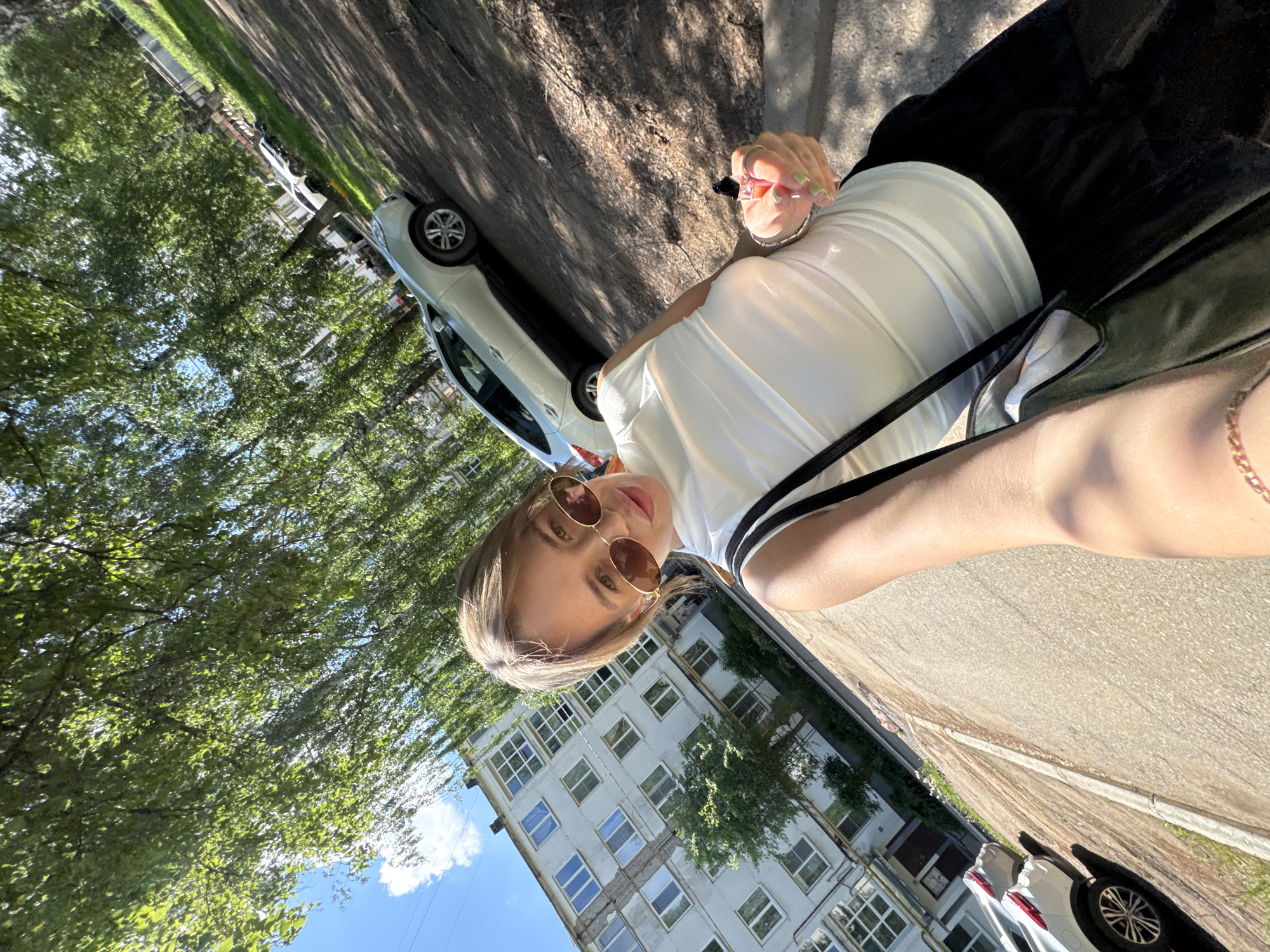 A woman wearing round sunglasses and a white top with black straps leans out of a car, holding a small, dark object. The location appears to be in Chaikovsky, Russia, characterized by multi-story, Soviet-era apartment buildings with many windows, visible in the background. Cars are parked along the street lined with trees and sunlight is prominent. The perspective is from a low angle.