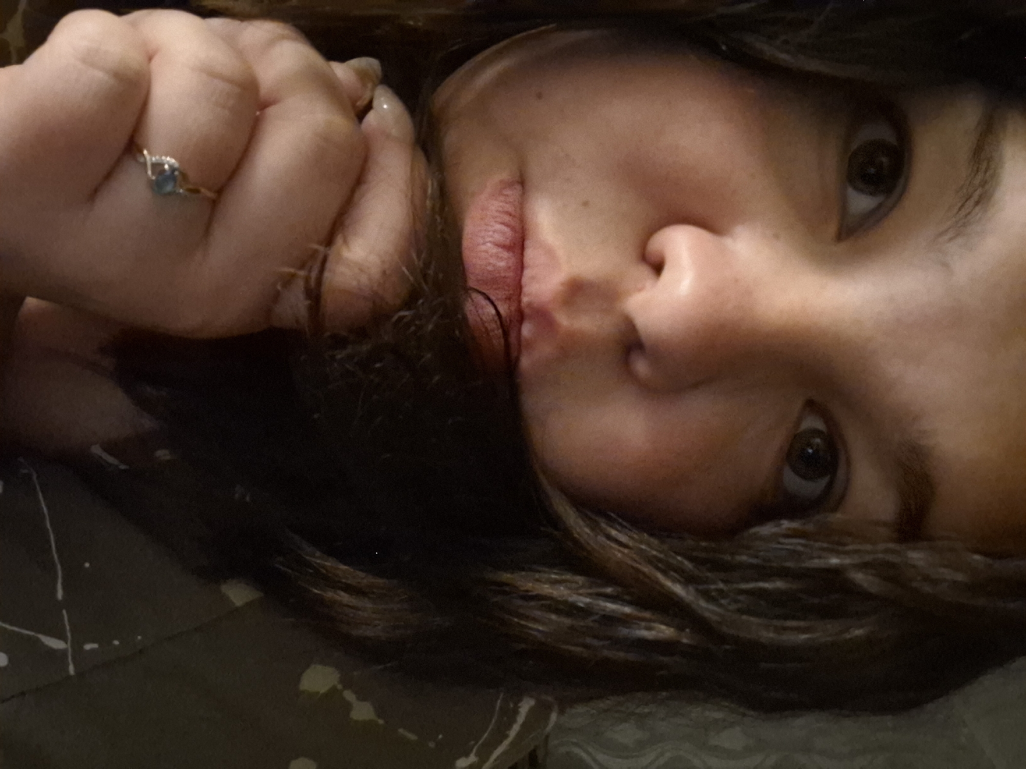 A close-up portrait shows a person with dark, slightly wavy hair, looking directly at the viewer with one wide-open eye. The head is tilted, giving a dynamic, almost reclined perspective. The person's lips are slightly parted, and their hand is positioned near their face, resting on or near their hair. On one finger of the hand, a silver-toned ring with a small light blue or green square-shaped gemstone, flanked by tiny white accents, is clearly visible. The skin tone is warm, and natural facial features are prominent. The background is a muted, dark fabric, possibly olive green or dark gray, adorned with irregular white and light yellow or green patterns that resemble splatters, suggesting a blanket or bedding. The lighting appears soft and ambient. No text is visible in the image, and there are no specific visual cues to indicate the provided location context of "Марковский, Russia." The overall impression is one of a direct, intimate, and perhaps curious gaze.