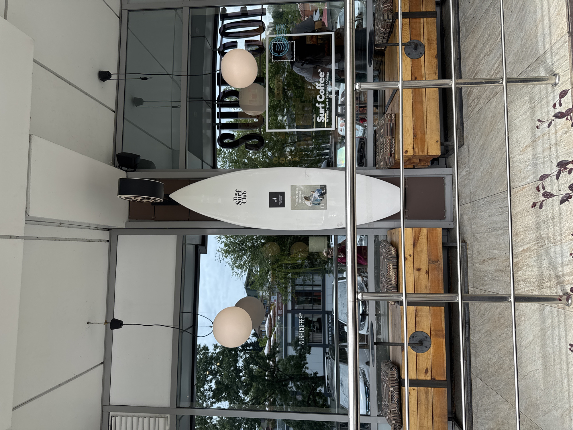 The image shows the exterior of a Surf Coffee establishment in Sochi, Russia. A white surfboard stands vertically in front of a large glass window. Inside the establishment, a person with a backpack is visible. The cafe features outdoor seating with wooden benches and patterned cushions. The window reflects trees, buildings, and cars, suggesting an urban environment. A sign on the window reads "Surf Coffee" with Russian text below it. The overall scene depicts a contemporary cafe with a surf-themed ambiance.