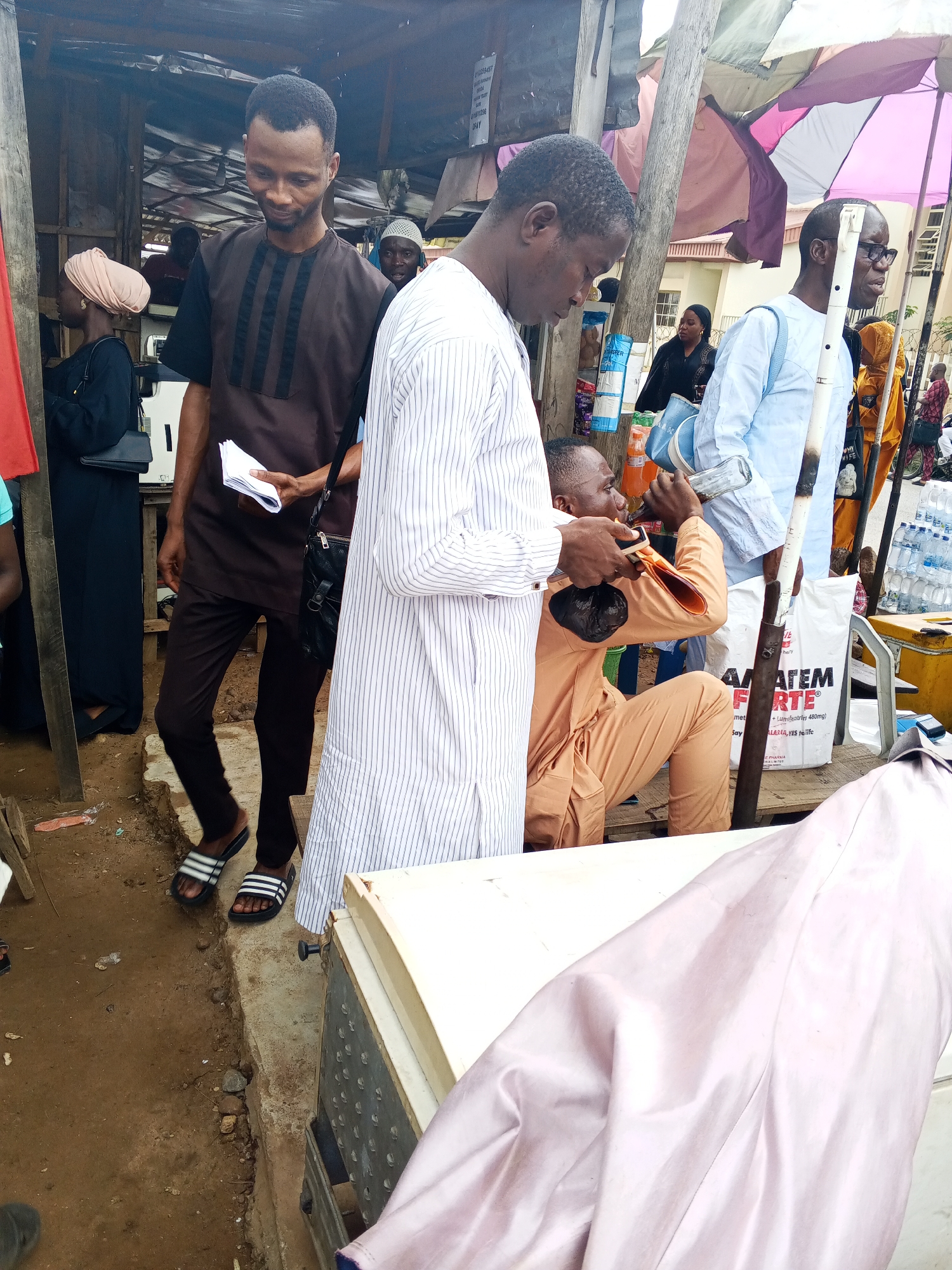 The image depicts a street scene in Lokoja, Nigeria, likely a market area. A group of individuals are gathered, some appearing to be vendors and others customers. In the foreground, a man in a brown and black dashiki is holding a stack of papers. To his right, another man in a light blue shirt is looking down. Further back, a person in an orange outfit is crouched down, holding a bottle to their mouth. Several other people are visible in the background, including women wearing headscarves and men in traditional attire. The environment appears somewhat cluttered, with stalls, umbrellas, and various items on display. The ground is dirt and concrete, suggesting an outdoor commercial setting.