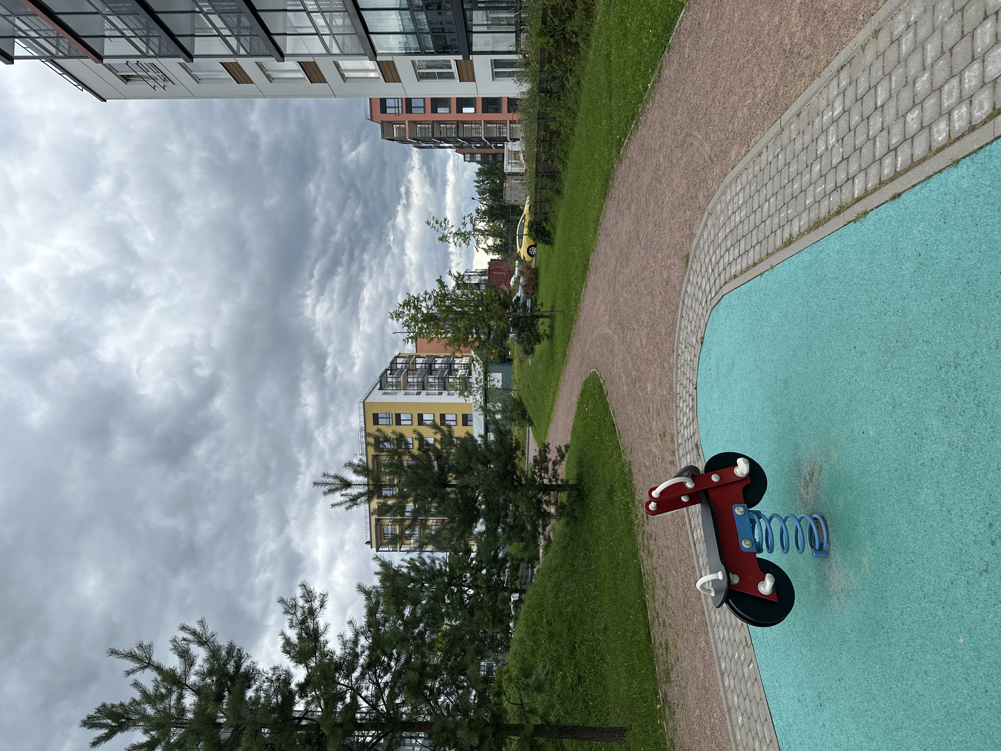 A playground scene is captured on an overcast day in the city of Vsevolozhsk, Russia. In the foreground, a red and black spring rider shaped like a motorcycle is situated on a bright teal rubber safety surface. The playground area is bordered by a walkway paved with grey interlocking bricks and a surrounding path made of reddish-brown gravel. Beyond the playground, a grassy hill slopes upward, dotted with young pine trees. In the middle ground, apartment buildings with modern architecture and balconies are visible. One building is yellow with orange accents, another is coral, and a large building on the right is white with dark-tinted glass balconies. A bright yellow car is parked near a row of trees and a construction fence in the distance. The sky is filled with heavy, grey clouds, suggesting an imminent change in weather.