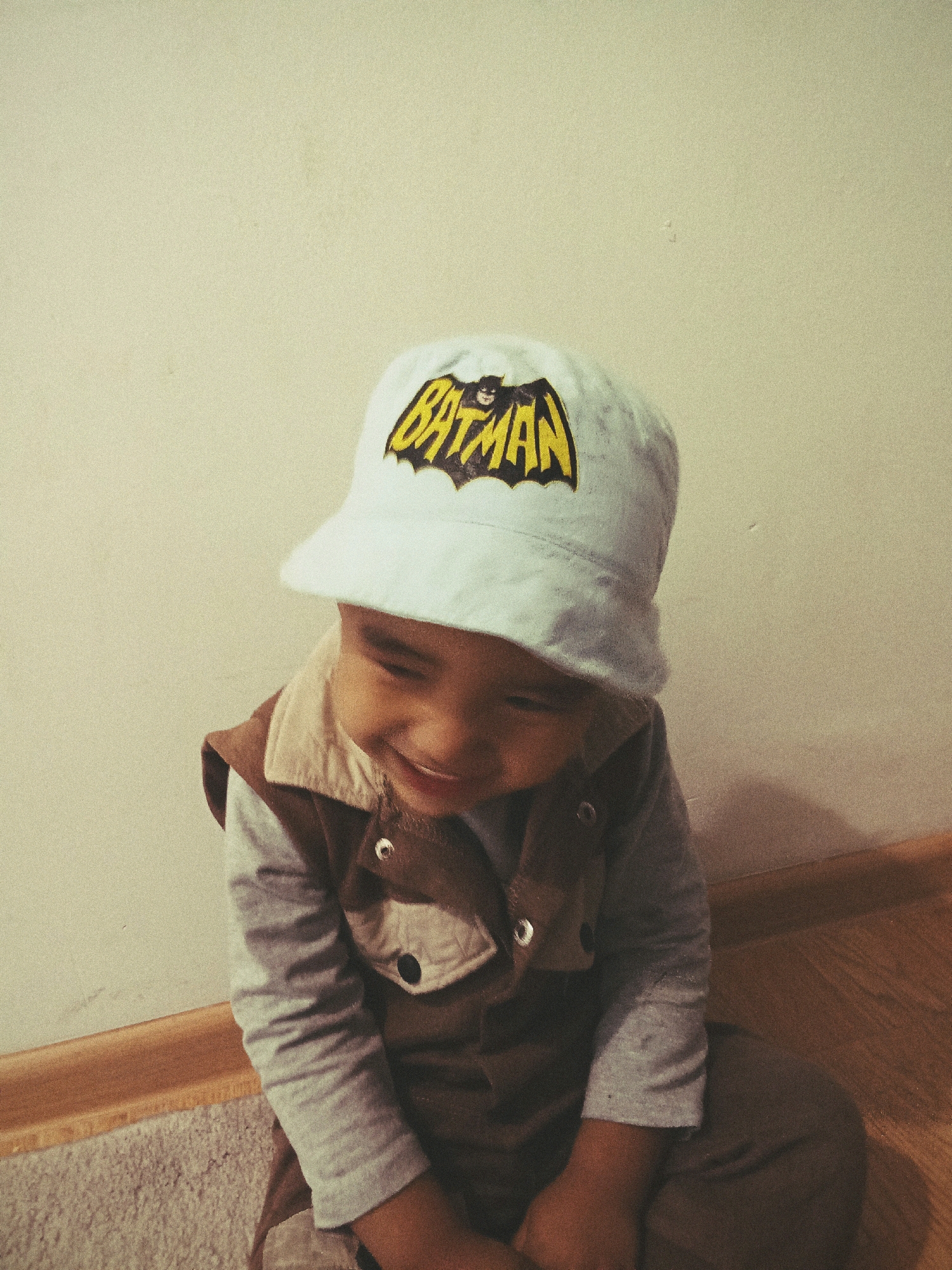 An undated, medium-shot photograph captures a young child wearing a white bucket hat with a yellow and black Batman logo. The child is also wearing a light gray long-sleeved shirt and a brown vest with a light-colored collar. The child is looking down and to the left with a slight smile. The background consists of a textured, light-colored wall and a wooden floor with a baseboard. The image was taken indoors, and there is no indication of the city of Astana, Kazakhstan, or any specific location.