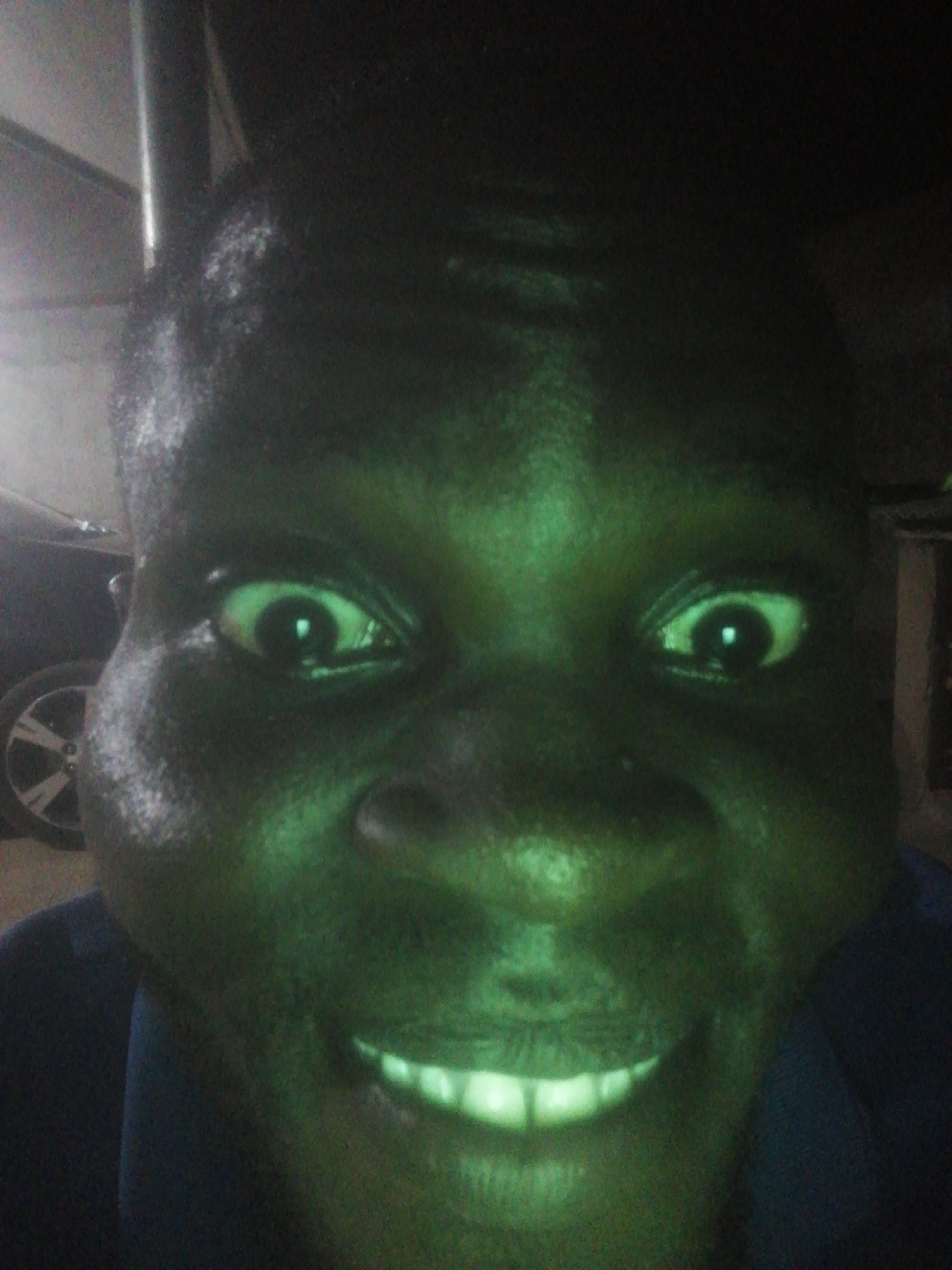 A close-up photograph shows a dark-skinned person with large eyes and a wide smile, illuminated by a green light source. Behind the person, on the left side of the frame, is a blurred image of a vehicle wheel. The background is dark and indistinct, making it difficult to discern specific details about the location, but given the prompt, the image was supposedly taken in Ikorodu, Nigeria.
