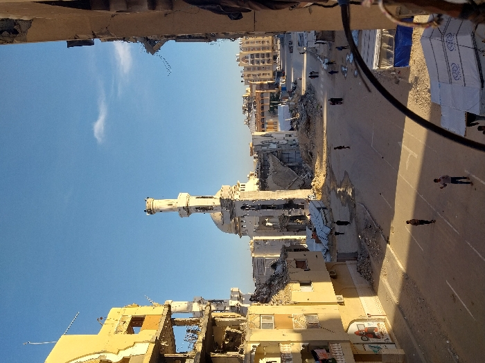 A street scene shows extensive damage to buildings, including a mosque with a prominent minaret. Rubble and debris are scattered among the damaged structures. Several individuals are walking or standing on the street, possibly surveying the destruction. Tents are visible at the right of the road, which might indicate temporary shelters. The overall scene suggests a post-conflict or disaster situation. The sky is clear and blue.