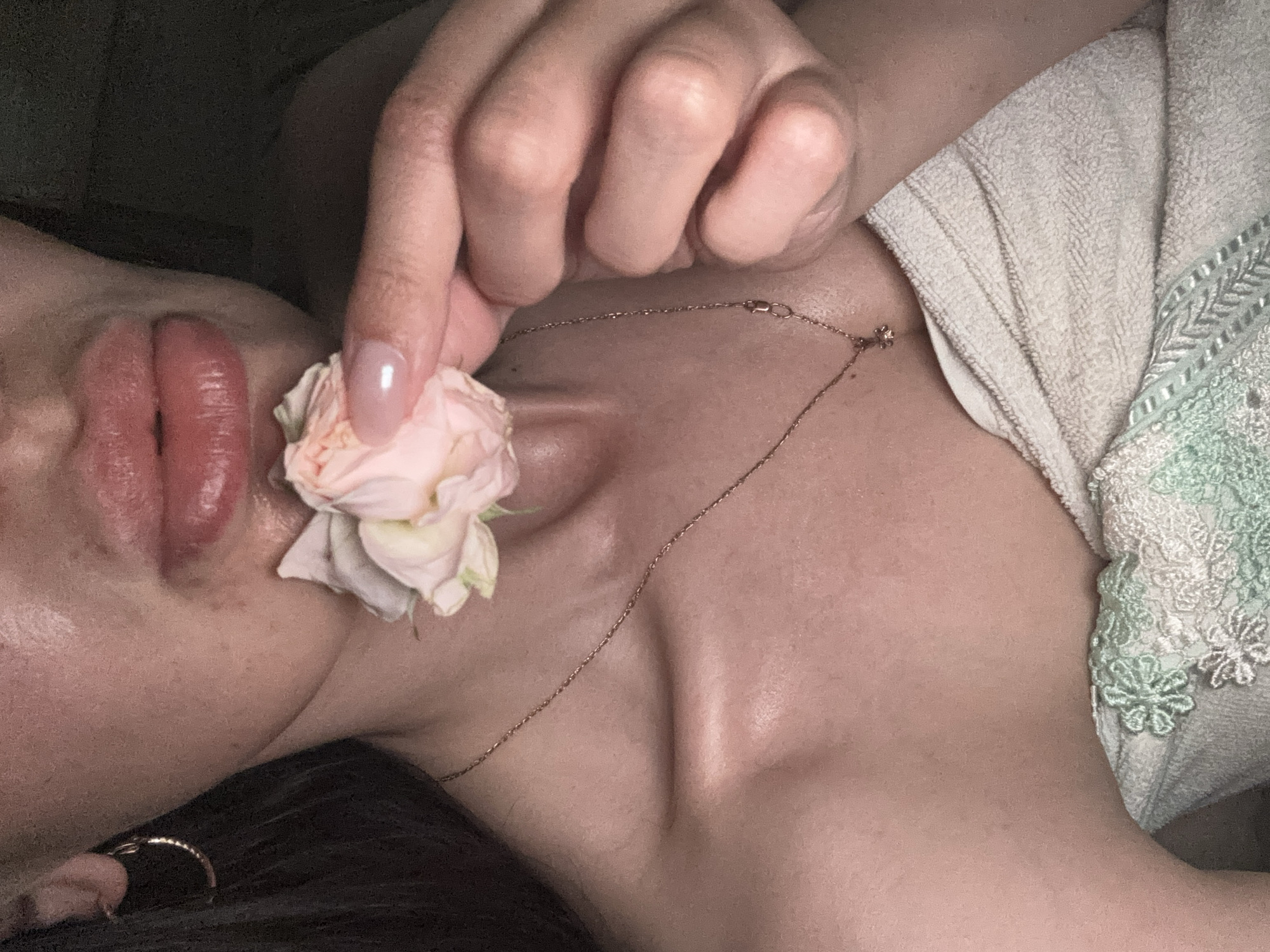 A close-up, top-down shot features a person, likely female, in a relaxed, intimate setting. The view focuses on their face from the mouth and chin downwards, extending to the upper chest and shoulders, with a hand occupying the upper center.
The subject's lips are full, reddish-pink, and appear moist or glossy, slightly parted. A manicured hand, adorned with a light, natural-toned nail polish, delicately holds a small, light pink rose to the side of the mouth and chin. A thin, gold or rose gold chain necklace with a small, subtle pendant is visible around the neck and resting on the collarbone. A simple ring can be partially seen on a finger at the bottom left of the frame.
To the right, a soft, light-colored fabric, possibly a towel or robe, is draped, showcasing decorative green and white floral embroidery or lace along its edge. The lighting is soft and warm, creating gentle highlights on the skin and enhancing a sense of tranquility. The overall impression is one of tenderness and quiet contemplation. No text is visible in the image. While the location context is provided as Sochi, Russia, there are no specific visual cues within the image to confirm this.