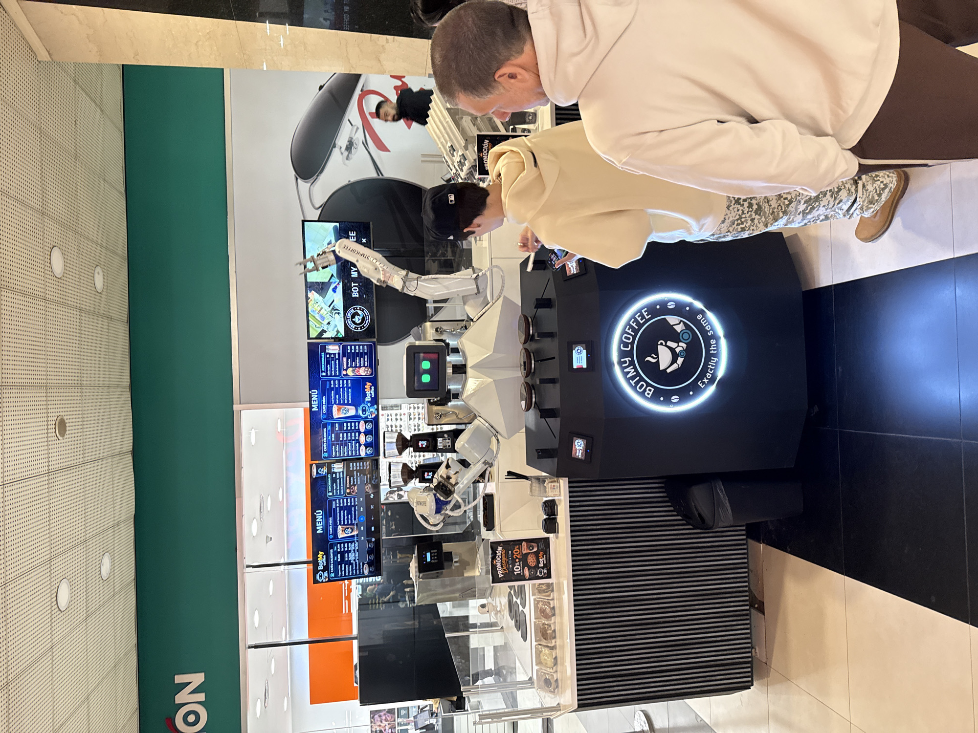 The image captures two men interacting with a "BotMy Coffee" robotic coffee station in a brightly lit indoor setting, likely a shopping mall in Buenos Aires, Argentina. A man in a light-colored hoodie and dark baseball cap leans forward, appearing to operate a touchscreen or observe the coffee preparation process. Beside him, another man in a white hoodie looks down at the machine.
The "BotMy Coffee" station features a prominent robotic arm, which is the central element for preparing beverages. Multiple display screens are visible, showing "MENÚ" (menu) options in Spanish, detailing various types of coffee and their prices. One screen also highlights a "PROMOCIÓN" (promotion) offering "10%-20%" off. The brand's circular logo, displaying "BOTMY COFFEE" and the tagline "Exactly the same" alongside a coffee cup icon, is visible on the side of the main unit.
The overall environment suggests a modern, functional space. In the background, a green wall with the partial text "NO" can be seen, indicating part of a larger sign. The floor has patterned black and white tiles, while the ceiling features recessed lighting. The men are dressed in casual attire, with one wearing camouflage-patterned trousers and tan boots. The scene depicts a modern automated service experience.