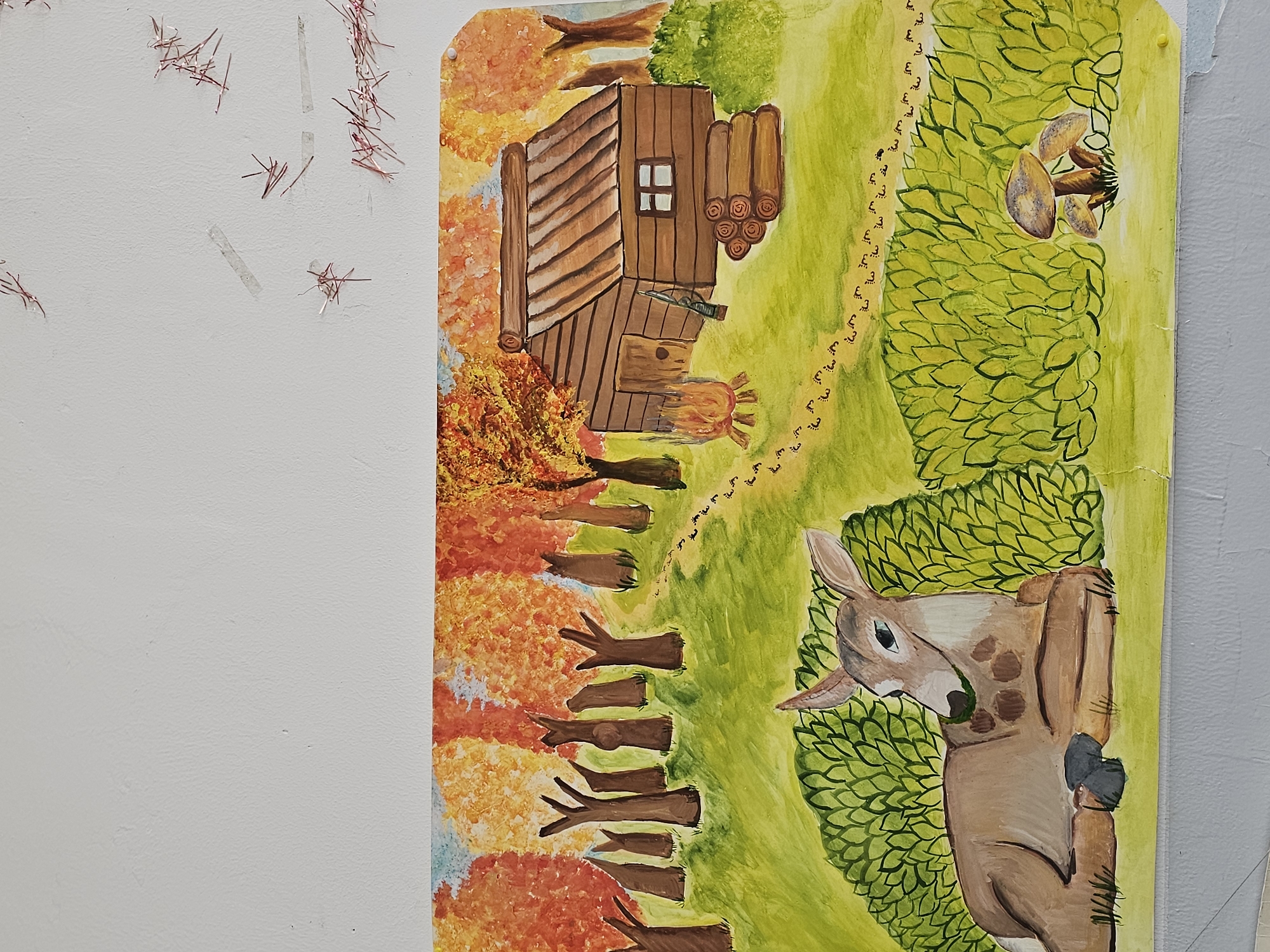 The image is a close-up, vertical shot of a hand-painted illustration. The scene depicts a woodland setting with a log cabin on the left side. The cabin is brown with a visible window, a stack of logs behind it, and what appears to be a rifle and a fire pit. Autumn trees with orange foliage surround the cabin. A path leads away from the cabin, marked by a trail of what appear to be footprints or small symbols. On the right side of the image is a deer partially hidden behind green foliage. The artwork is attached to a white wall with some visible red threads or fibers. The background surrounding the artwork is plain and undetailed. Yerevan, Armenia, is not depicted in the image. There is no evidence that Yerevan, Armenia, is the location.