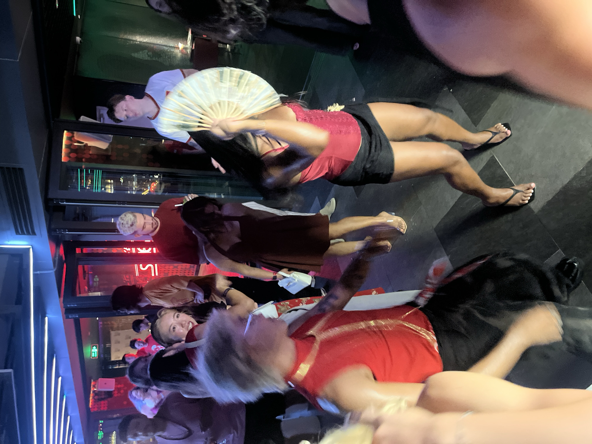The image shows a group of people at a party, possibly in a club or bar, in Đà Nẵng, Vietnam. The lighting is dim with neon blue and red accents, suggesting it's nighttime. Several individuals are visible, some in focus and others blurred due to movement.
In the foreground, a person with blonde hair wearing a red outfit is blurry, and their face is partially obscured. To their right, another person's legs and feet are visible, wearing flip-flops.
Further back, a woman in a red top and black skirt is holding a large, light-colored fan, obscuring her face. She is standing and appears to be posing. Beside her, a person in a brown dress is also visible. In the background, a man wearing a white t-shirt with red trim is looking towards the camera, holding a fan. Other people are present, some smiling and looking at the camera.
The setting appears to be indoors, with a dark floor and a background that suggests a venue with windows looking out onto a city scene, possibly with illuminated buildings. The overall atmosphere seems to be lively and celebratory. There is no visible text in the image.