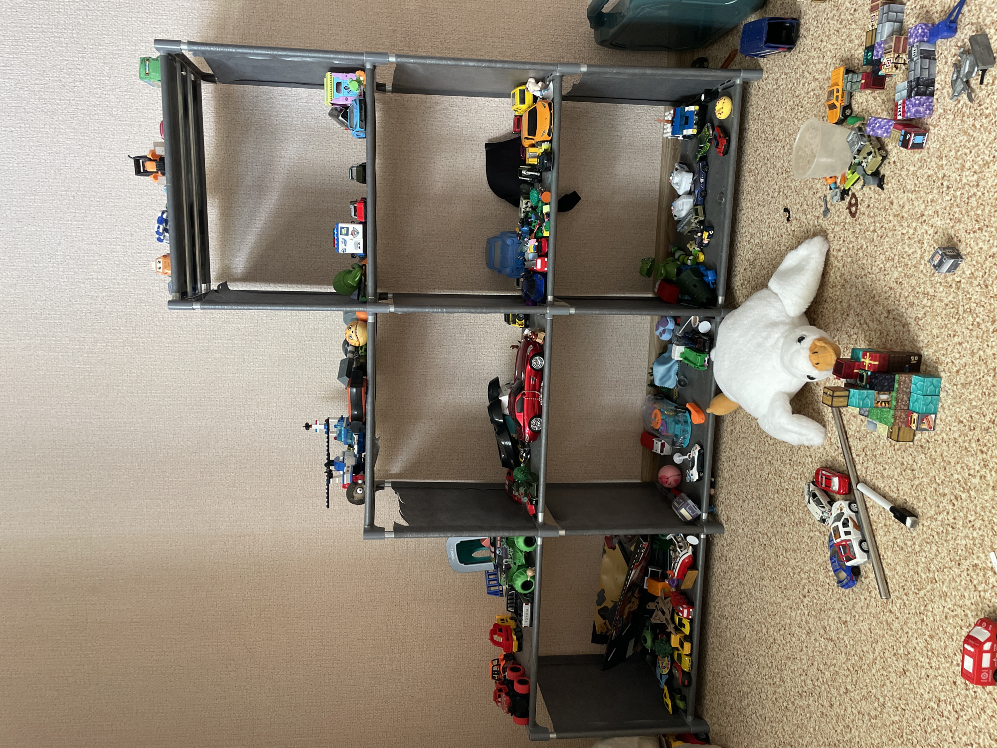 A tall, grey fabric shelf unit filled with toy cars and building blocks stands on a beige carpeted floor. The shelf unit is divided into multiple compartments, each containing a variety of toys.
On the top shelf, on the left side, is a green toy truck. To its right, a blue toy car and a yellow toy car are visible. Further to the right, a black bag lies on the shelf.
The second shelf from the top, on the left, has a red toy car and a white and blue toy car with a ladder. On the right side of this shelf, there are numerous toy cars, including a yellow sports car, an orange car, and a black car. A large white plush toy with orange accents is lying on the carpet next to the shelf.
The third shelf from the top displays a red sports car and a black car on the left, and a blue toy truck and other small toys on the right.
The bottom shelf, on the left, contains a green toy truck, a blue toy car, and several other small toy cars. On the right side of this shelf, a collection of colorful building blocks resembling Minecraft blocks are arranged, along with more toy cars and a white pen.
In the background, out of focus, a large dark green plastic container can be seen on the floor. The scene appears to be indoors, in a domestic setting. There is no indication of the time of day or weather conditions. The overall impression is a child's play area with a large collection of toys.