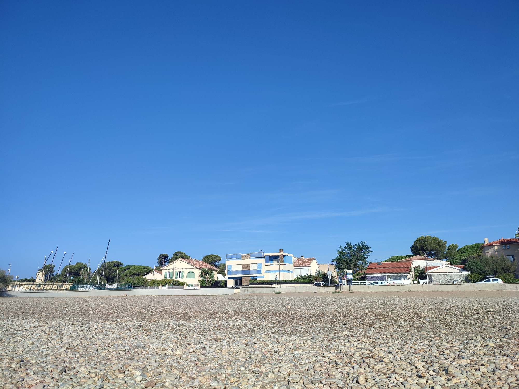 The image depicts a bright, sunny day with a clear blue sky over a pebbled beach. In the background, a row of houses and buildings are visible, lining the coast. Some of these structures appear to be residential, with red-tiled roofs and windows. Among the buildings, there are also several tall masts, suggesting the presence of a marina or sailing club. Trees and greenery are interspersed between the buildings and behind them, indicating a coastal town or resort area. A low concrete wall separates the beach from the built-up area. A few cars are parked along the road behind the wall. The foreground is dominated by the textured surface of the beach, composed of numerous small, smooth pebbles in various shades of gray, brown, and tan. The overall scene conveys a sense of a peaceful, seaside environment, likely a popular spot for leisure activities. This scene is consistent with the coastal areas of Hyères, France, known for its beaches and harbors.