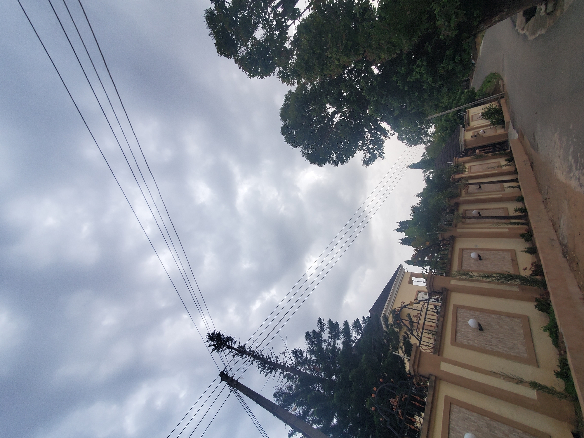 An exterior, low-angle shot captures a residential fence and road on a cloudy day in Jos, Nigeria. The fence, painted in shades of beige and light brown, features decorative panels and ornate metalwork along the top. Behind the fence, a light yellow house with a dark roof is partially visible. Tall evergreen trees and other foliage are scattered throughout the scene. Overhead, multiple power lines stretch across the sky, supported by a concrete pole on the left side of the frame. A paved road curves into the distance on the right, bordered by a large tree with dark green leaves. The sky is overcast with a uniform layer of gray clouds. There are no people visible in the image.