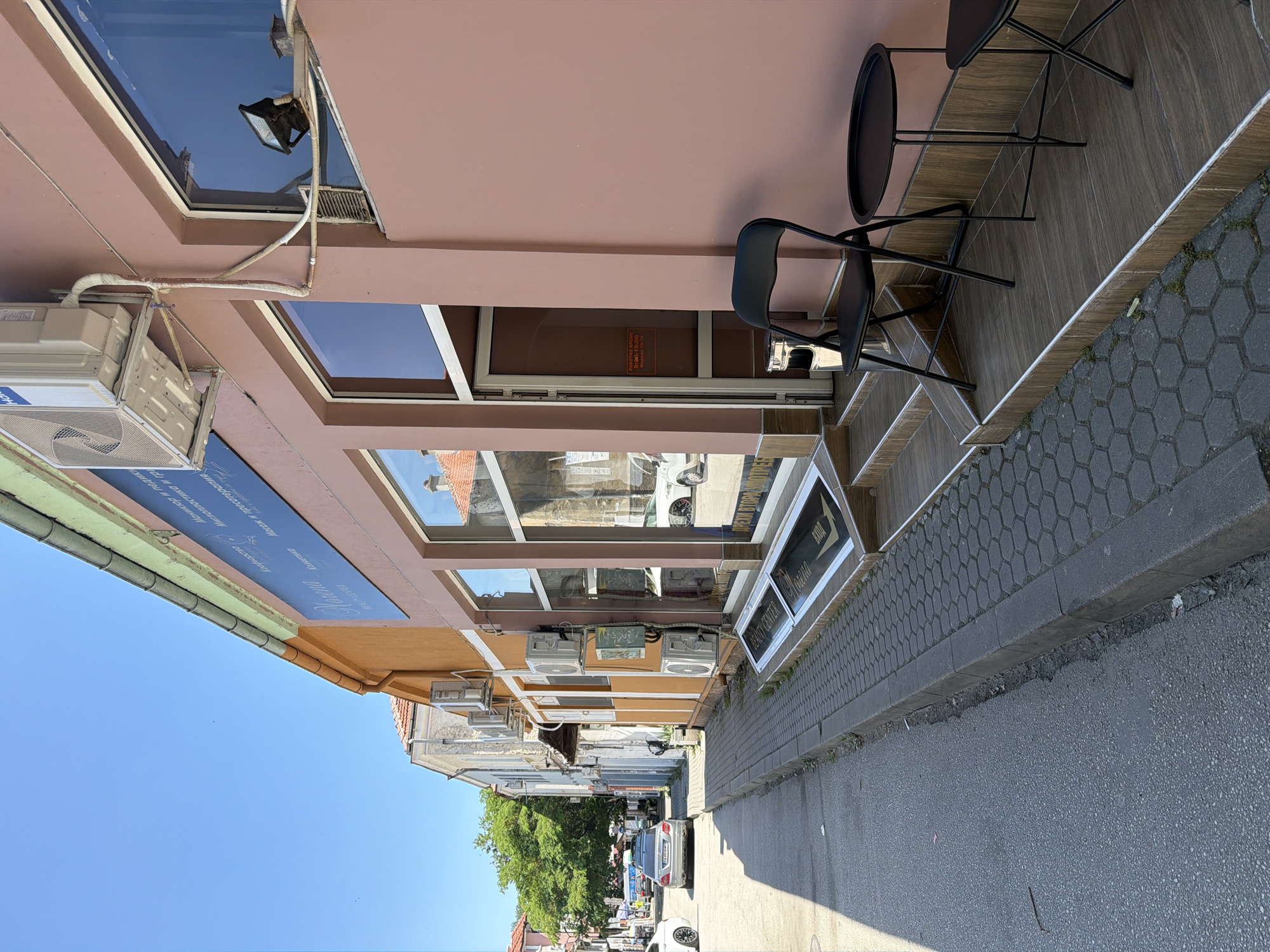 The image depicts a street view in Pazardzhik, Bulgaria. The main subject is the facade of a building with a pinkish-brown exterior. The building features a storefront with large glass windows and a prominent entrance. Several steps lead up to the entrance, and there is outdoor seating consisting of a black folding chair and a small black table. An air conditioning unit is visible mounted on the exterior wall above the storefront.
To the left of the building, a sidewalk paved with hexagonal tiles runs alongside the street. The street itself is paved with asphalt and shows some wear and tear. Several cars are parked or driving on the street, including a silver sedan and a white van. Adjacent buildings of varying colors, including an orange one, line the street, suggesting a commercial or mixed-use area. Green trees are visible in the background, indicating an urban setting with some vegetation. The sky is clear and blue, suggesting a sunny day.
A sign with text in Cyrillic script is mounted on the building above the storefront. Another sign, also in Cyrillic, is affixed to the glass door of the entrance. A small, silver trash receptacle is positioned near the entrance. The overall impression is that of a typical street scene in a Bulgarian city.