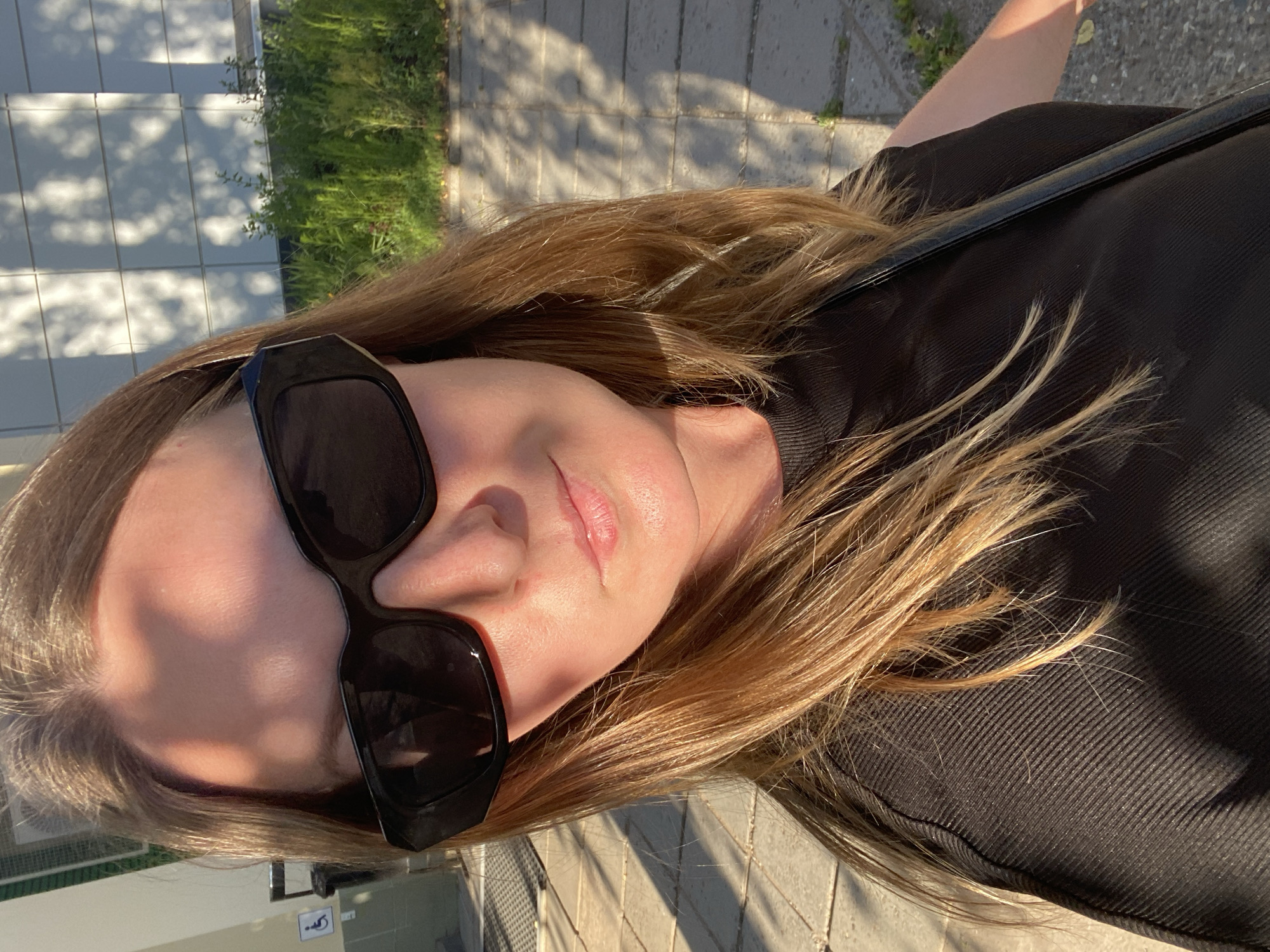A woman with long, blonde hair and sunglasses is captured in a selfie. She is wearing a black top and a strap from a bag crosses her shoulder. The image is taken outdoors with dappled sunlight casting shadows on the ground, suggesting it is either morning or late afternoon. In the background, there is a building with a tiled facade and some greenery. The overall impression is casual and relaxed, possibly taken while walking or enjoying a sunny day. There is no visible text in the image.