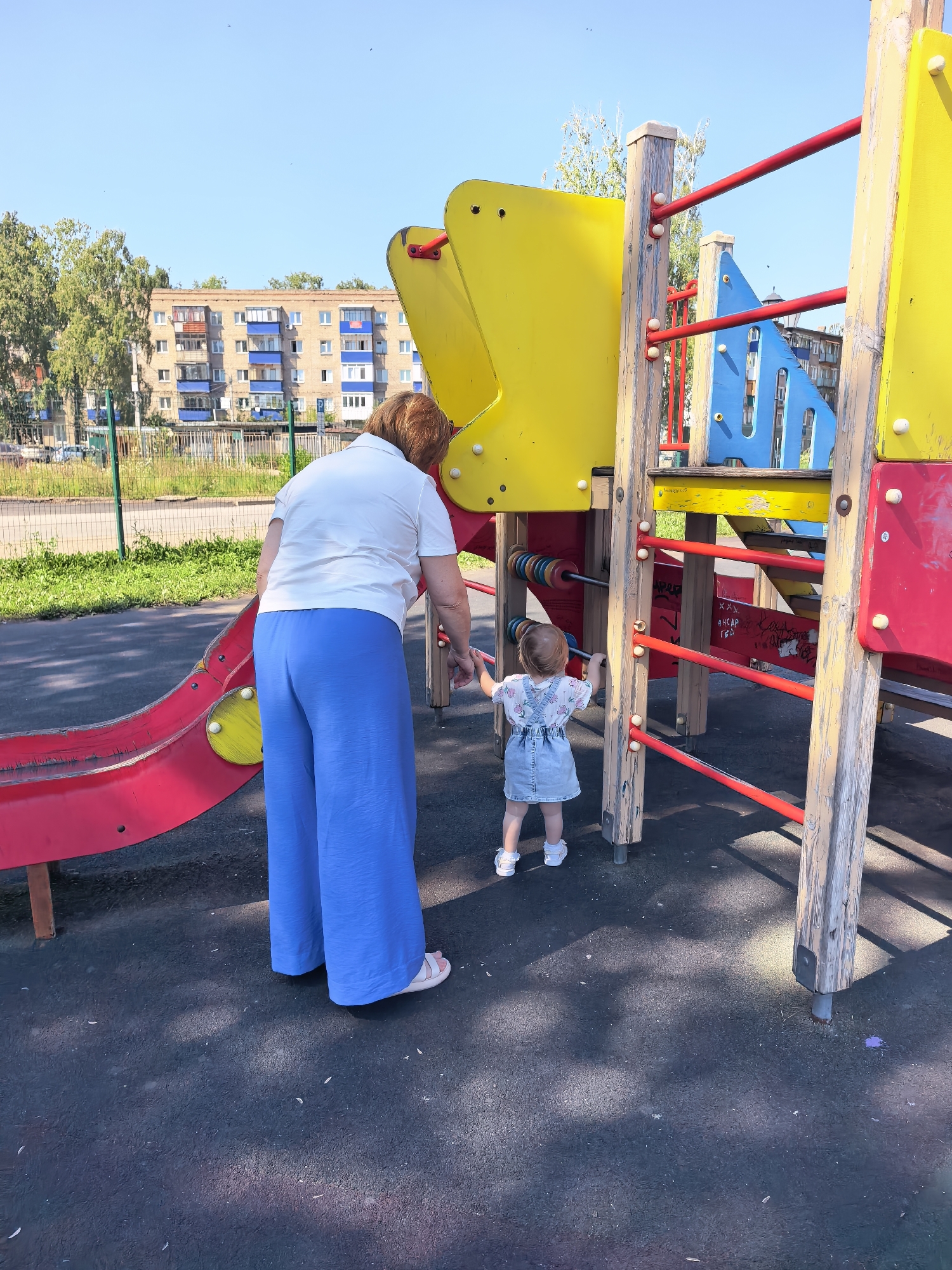 The image depicts a playground with a woman and a young child on a sunny day. The woman, wearing a white polo shirt and blue wide-leg pants, stands with her back to the camera, looking towards a playground structure. The child, in a denim dress and white shoes, faces the structure, touching its wooden posts. The playground features colorful sections of yellow, red, and blue, with red bars and wooden supports. An abacus with colorful beads is attached to the structure. In the background, apartment buildings and trees are visible, suggesting an urban environment. The ground is a dark, textured material, likely rubber safety surfacing common in playgrounds. The sky is clear and blue. The location is identified as Sterlitamak, Russia, based on context provided.