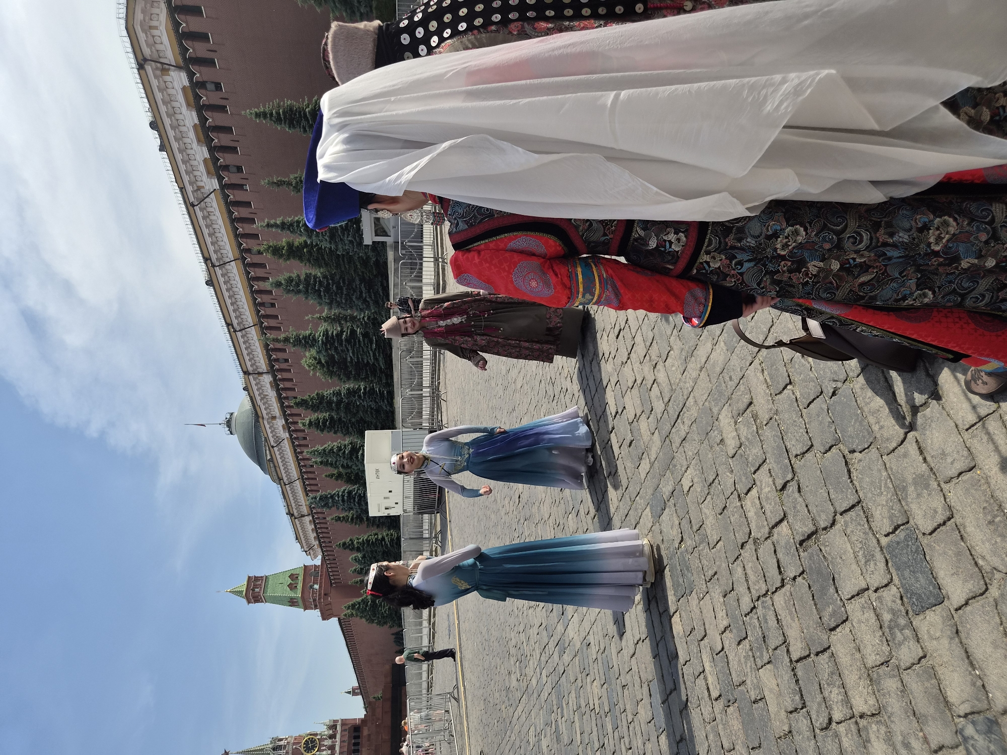 This image captures a bright outdoor scene in Moscow, Russia, specifically Red Square, with the Kremlin Wall and towers forming the background under a clear blue sky. The foreground consists of rough, grey cobblestone paving.
On the right side of the foreground, two individuals are seen from behind, dressed in elaborate traditional garments. The person closer to the viewer wears a richly embroidered dark garment with red sleeves and intricate floral patterns, along with a blue hat and a large white fabric draped over their back and head. To their right, another individual wears a dark garment adorned with numerous silver or white circular embellishments and a light brown fur hat.
In the mid-ground, slightly to the left, two women face each other, both wearing long, flowing, ombre dresses that transition from dark blue/teal at the top to light blue/white at the hem. These dresses feature long sleeves and subtle embellishments. Both women wear distinctive headpieces. The woman on the left has her back partially to the camera, looking towards the woman on the right, who smiles slightly towards the viewer.
Further back, behind these women, a white rectangular utility box labeled "ИНТЕХ" (INTECH) is visible, positioned in front of a row of small, conical evergreen trees. Metal crowd control barriers stretch across the mid-ground. To the far left, a man in a green shirt and dark pants stands behind these barriers. To the right of the women in blue, another woman in traditional attire, featuring a brown long-sleeved dress with multiple necklaces and a light brown hat, walks towards the group in the foreground.
The background is dominated by the historic red brick Kremlin Wall with its distinctive battlements. Visible towers include the Spasskaya Tower on the far left, featuring a clock and a red star at its spire, and a tower with a green pyramidal roof further right. The green dome of the Senate Palace is also visible behind the wall. The scene is brightly lit by sunlight, casting distinct shadows on the cobblestones.