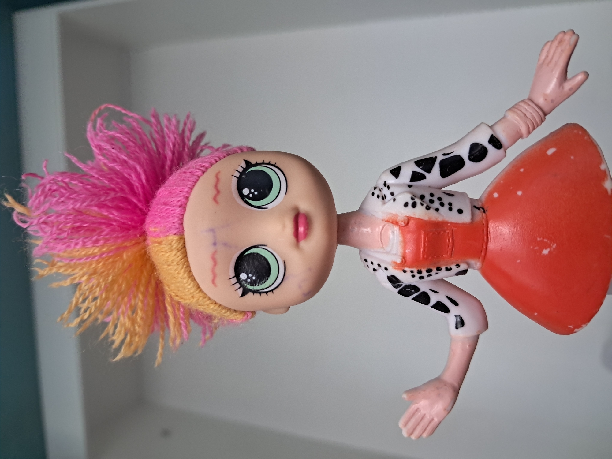 A close-up, eye-level, indoor studio shot shows a doll against a light gray background with a hint of a teal ceiling at the top. The doll is a fashion doll with a light skin tone and a punk-inspired appearance. It has large, green eyes with prominent black eyelashes and eyebrows drawn with squiggly lines. Its lips are painted pink. The doll's hair is styled in a mohawk shape, with the left side blonde and the right side bright pink. It is wearing an orange dress with a cinched waist and a flared skirt. Over the dress, it has a white jacket with black polka dots and triangular patterns. The doll's left arm is extended outward, palm facing up, while its right arm is bent at the elbow. The doll appears to be made of plastic, with some visible paint scuffs on its dress and jacket. The lighting is even, casting no significant shadows.