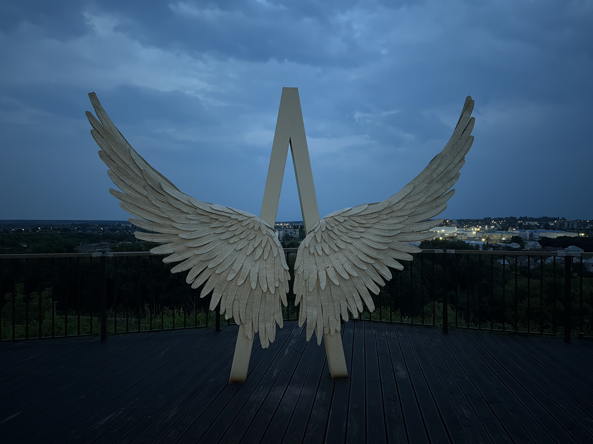 This is a medium shot, eye-level, outdoor, horizontal photograph taken at dusk. The foreground is a dark wooden deck with planks running horizontally. A black metal railing is visible behind the deck, with dark trees and a distant city skyline beyond. In the center of the frame, a large, stylized sculpture of a pair of white angel wings is mounted on a vertical, A-frame structure. The wings are symmetrical and appear to be made of layered, feather-like elements. The sky above is a deep, overcast blue with dramatic cloud formations. The city lights in the distance suggest an urban or suburban environment. The scene is lit by ambient twilight. The location is identified as the city of Lebedyan, Russia.