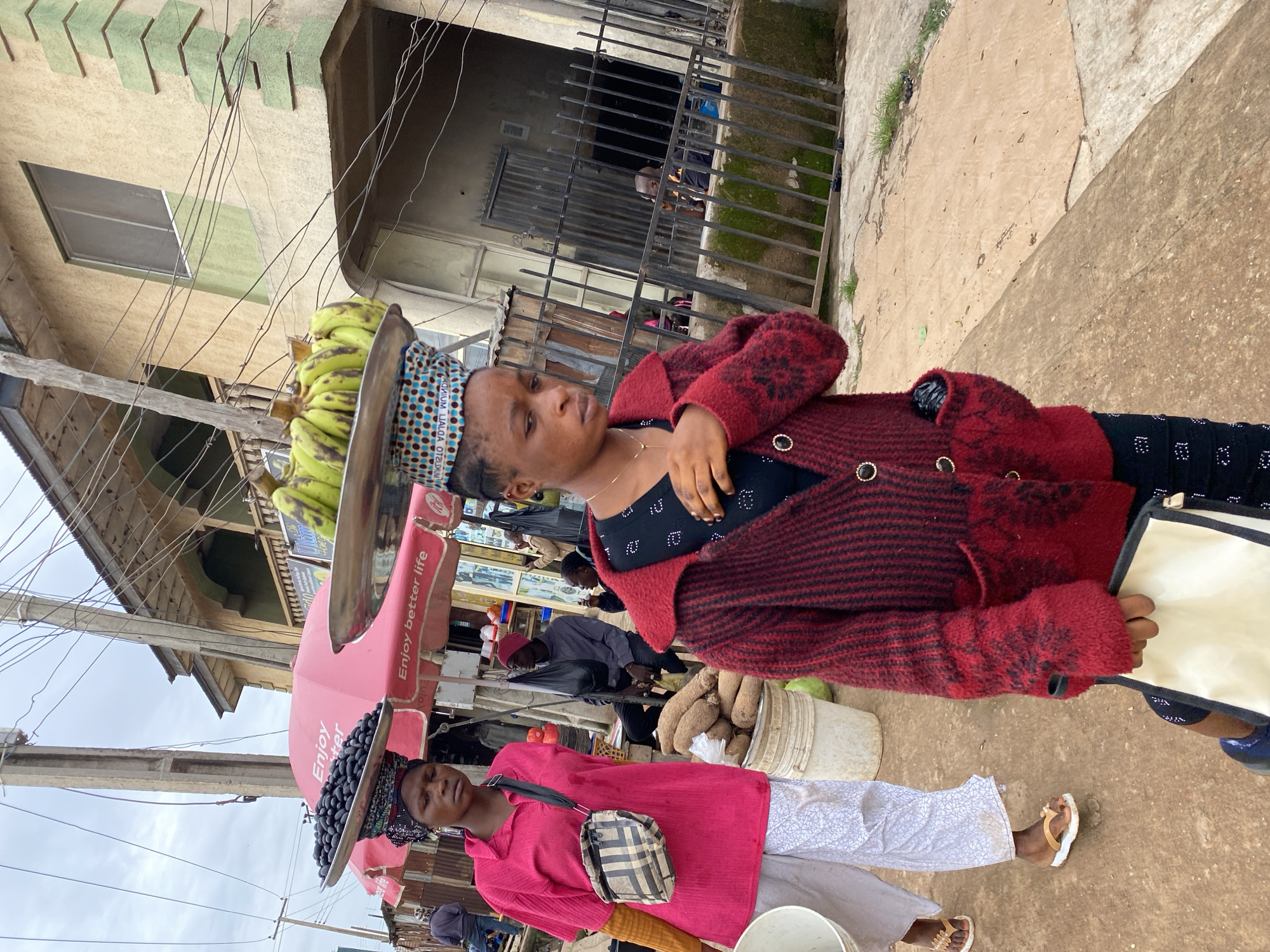 In Bukuru, Nigeria, two women are walking along a dusty street. The woman in the foreground is wearing a red and black patterned cardigan over a black top and patterned trousers. She is carrying a white pouch in her left hand and has a patterned cloth on her head, supporting a metal tray filled with green bananas. The woman behind her is wearing a bright pink collared shirt and light-colored patterned trousers. She has a black and beige patterned crossbody bag. On her head, she carries a metal tray laden with dark berries. Behind them, a stall with a pink umbrella displays "Enjoy better life" text. Several buckets, one white and one cream-colored, are visible, containing what appear to be yams. In the background, a multi-story building with cream-colored walls and green accents stands, featuring windows and a metal fence. Other individuals are present in the background, some sitting and others walking. Overhead, utility wires crisscross the sky.