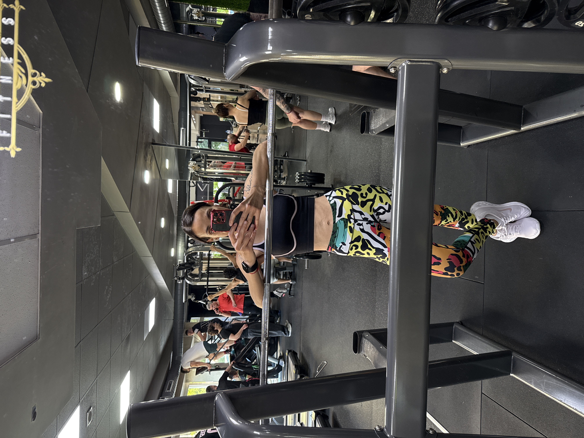 A woman wearing a black sports bra and patterned leggings is taking a selfie in front of a mirror in a gym. The gym has other people working out in the background, with various exercise machines and weights visible. The location appears to be a fitness center, possibly in Pleven, Bulgaria, based on the logo partially visible in the upper left.