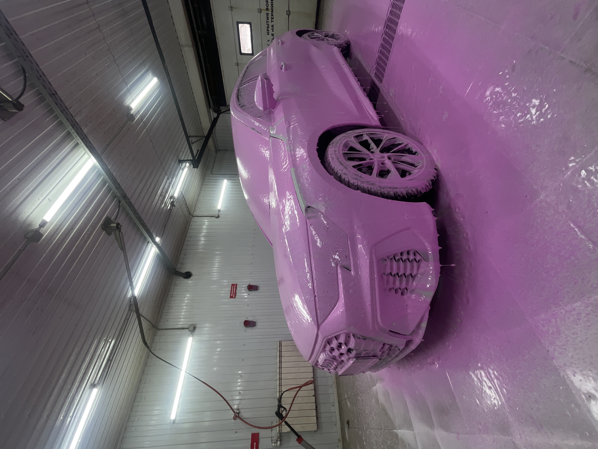 A car is covered in pink foam at a car wash in Krasnoyarsk, Russia. The building has fluorescent lights and metal walls. A hose and nozzle are visible on the left side of the frame.