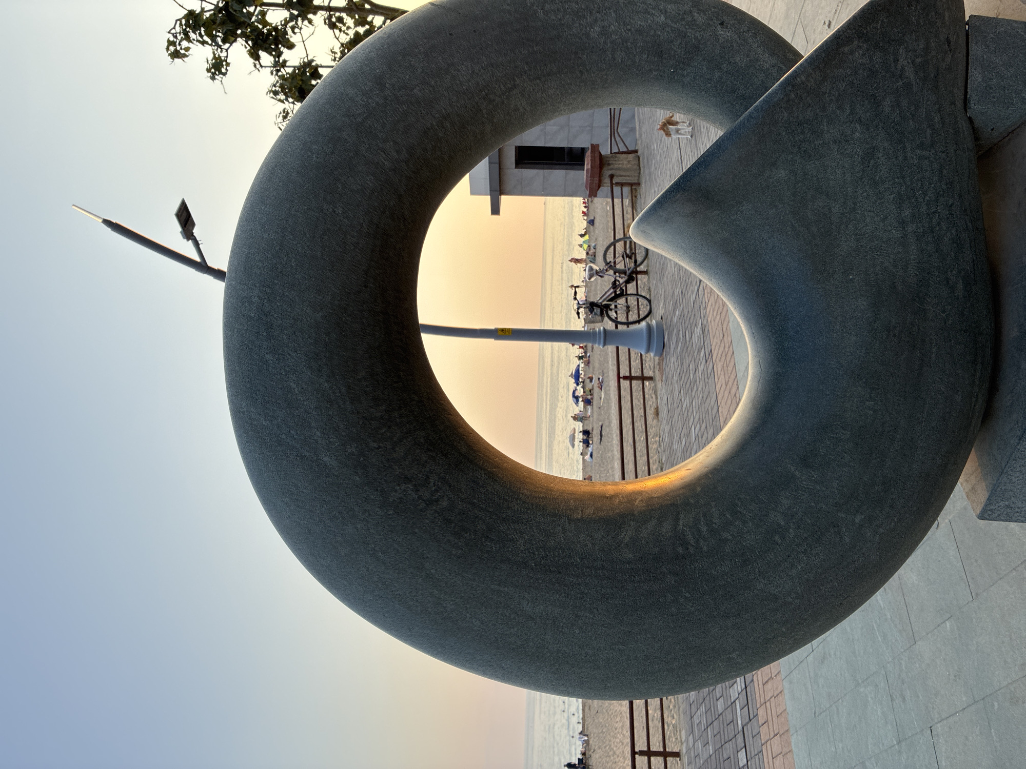 The image depicts a large, circular, gray stone sculpture in the foreground, with a portion of its opening framing a view of a beach and the sea at sunset. The sculpture has a rough, textured surface. The beach in the background is populated with several people sunbathing and sitting under umbrellas. A wooden railing separates the beach from a paved walkway where a bicycle is parked next to a lamppost. Further in the background, a small building is visible, and a tan and white dog is standing on the walkway. The sky is a soft gradient of pale blue and orange, indicative of sunset. The overall scene suggests a public area near a beach in Alanya, Turkey, likely a promenade or park.