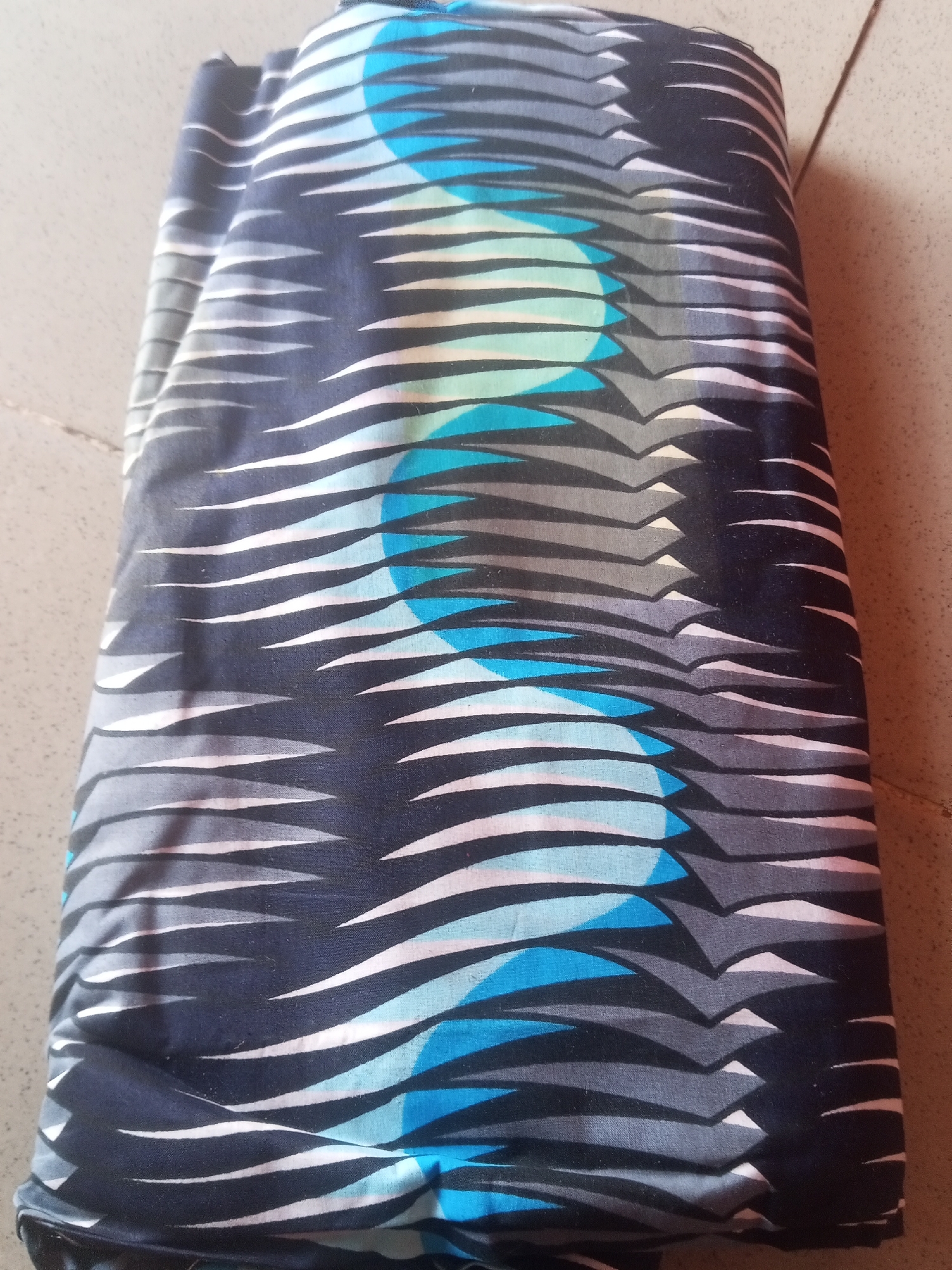 A rolled-up piece of fabric with a dark blue, grey, white, and light blue abstract pattern is lying on a tiled floor. The pattern consists of irregular wavy lines and geometric shapes that create a dynamic and somewhat chaotic visual effect. The fabric is folded lengthwise, with the pattern visible on the exterior. The lighting in the image is overhead and casts subtle shadows on the fabric and the floor. The background shows a light-colored tiled surface, likely a floor in an indoor setting. No people or specific actions are depicted.