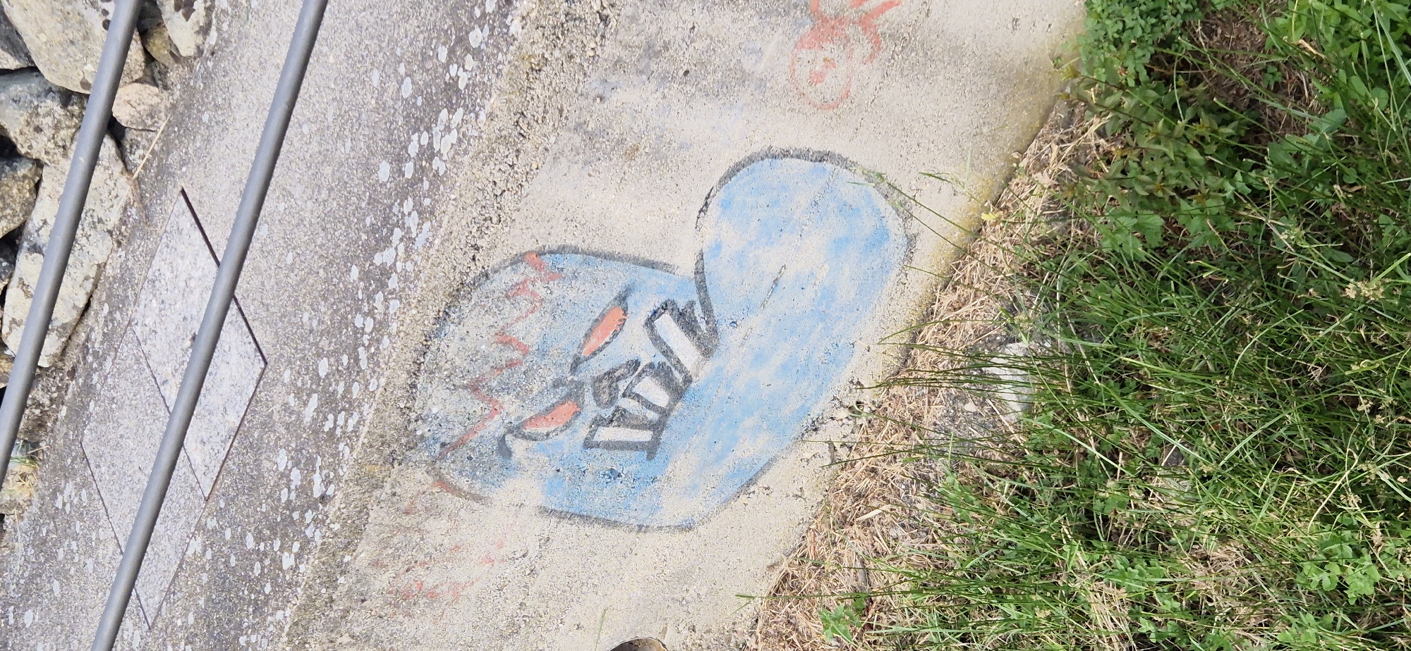 A close-up, eye-level shot shows graffiti on a concrete wall. The graffiti depicts a cartoonish, blue, blob-like creature with red, almond-shaped eyes and a jagged red line above its eyes, suggesting eyebrows or a furrowed brow. The creature has a wide, black outline around its face, and its mouth is open, revealing several white, angular teeth. The graffiti is stylized, with bold lines and vibrant colors, and appears to be spray-painted.
The concrete wall is weathered, with some patches of lighter discoloration and speckled textures. In the background, out of focus, a metal railing runs horizontally across the top of the image, separating the wall from a rocky embankment. To the left of the wall, there is a patch of dry grass and straw, and to the right, lush green grass and some small plants are visible, partially obscuring the base of the wall. The overall lighting suggests daylight. No people or active events are depicted. The location is identified as an outdoor urban environment, specifically on a concrete wall that might be part of a pedestrian pathway or retaining wall.