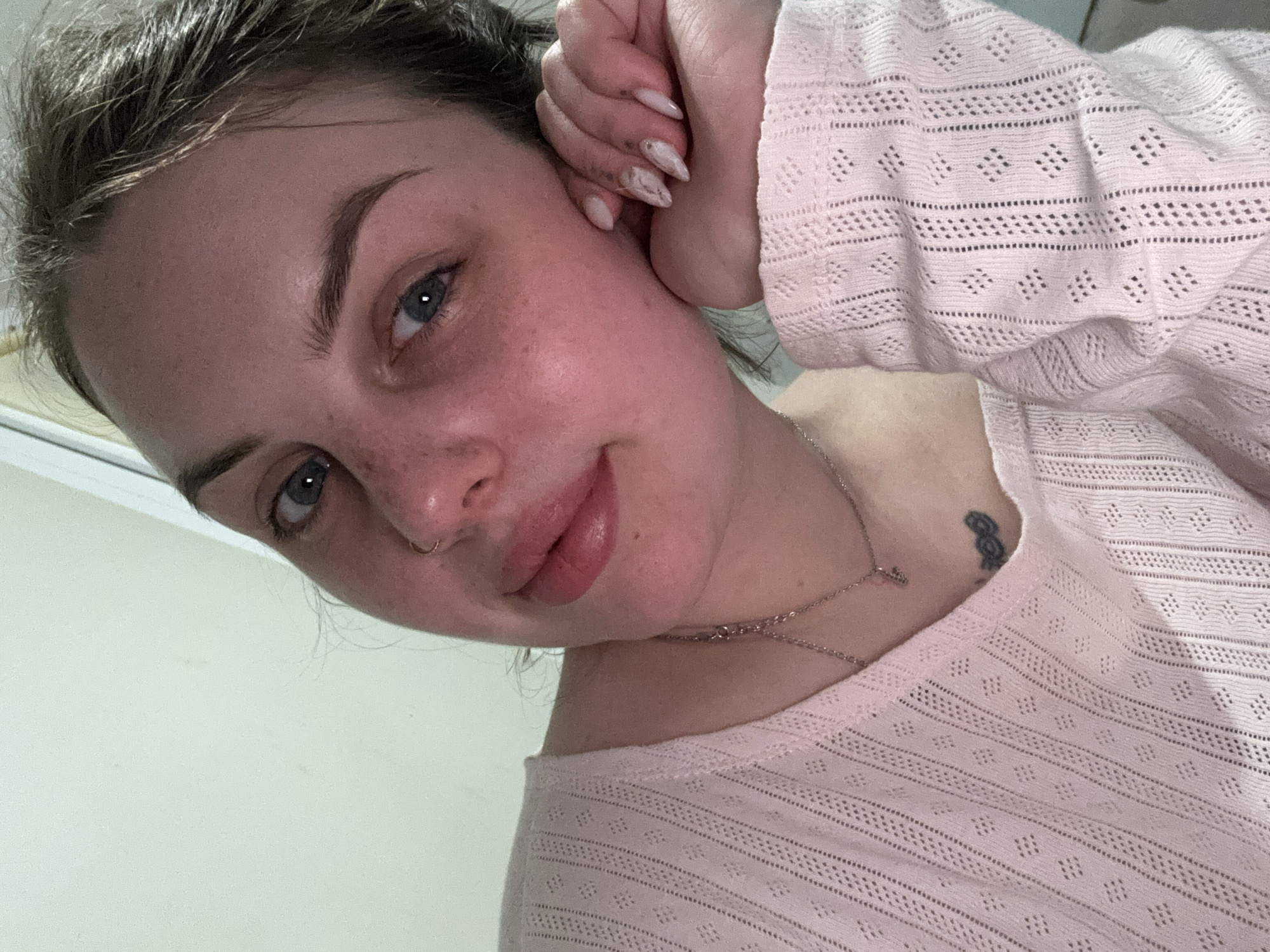 The image displays a young woman captured in an upward-angled close-up shot. Her head is gently tilted, and her right hand rests near her head, partially revealing nails with light-colored polish, possibly a marble or abstract design. She looks directly at the camera with blue eyes and a subtle, relaxed smile. Her fair complexion is dotted with freckles across her nose and cheeks. She wears a gold-colored hoop nose ring in her right nostril.
She is dressed in a light pink, long-sleeved top or sweater featuring a delicate open-knit pattern with small diamond shapes and horizontal lines of small holes. A thin silver chain necklace with a small, unidentifiable pendant is visible around her neck. On her left collarbone, a small, dark tattoo resembling the number "8" or an infinity symbol can be seen. Her hair, dark brown or light brown, appears to be casually styled, possibly pulled back.
The background is a plain, light-colored surface, indicative of an indoor setting, possibly a wall or ceiling. The lighting is soft and diffused. No specific location cues, time of day, or weather details are discernible from the image, nor is there any visible text. The overall expression is calm and composed.