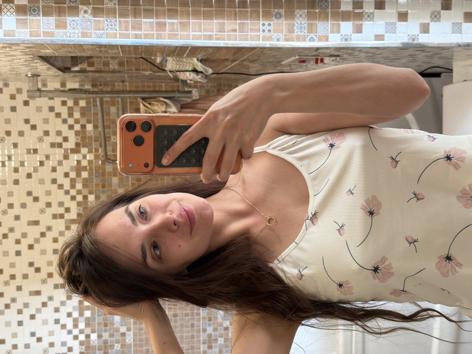 A young woman with long, dark, wavy hair is taking a selfie in a bathroom. She is wearing a white camisole with a floral pattern of pink flowers and green stems. She has a delicate gold necklace with a crescent moon pendant. She is holding her left hand up, touching her hair. Her right hand is holding an orange phone with a black case, and she is using it to take the picture in a mirror. The bathroom has tiled walls with a mosaic pattern of beige, brown, and white squares. There is a towel rack behind her, and a white toilet to her left. The lighting suggests it is daytime, and the overall impression is of a casual, private moment.