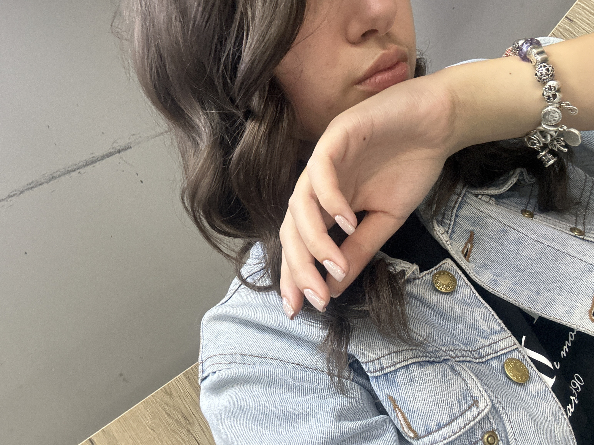 A close-up shot focuses on a person wearing a light-wash denim jacket with brass buttons. A Pandora-style bracelet is visible on their wrist. The person's hand is positioned near their face, with a partial view of their nose and lips. The fingernails are polished with a light color and subtle glitter accents. The background appears to be a plain, possibly neutral-colored wall above a wooden surface. No specific location in the city of Unknown, Unknown, can be determined.